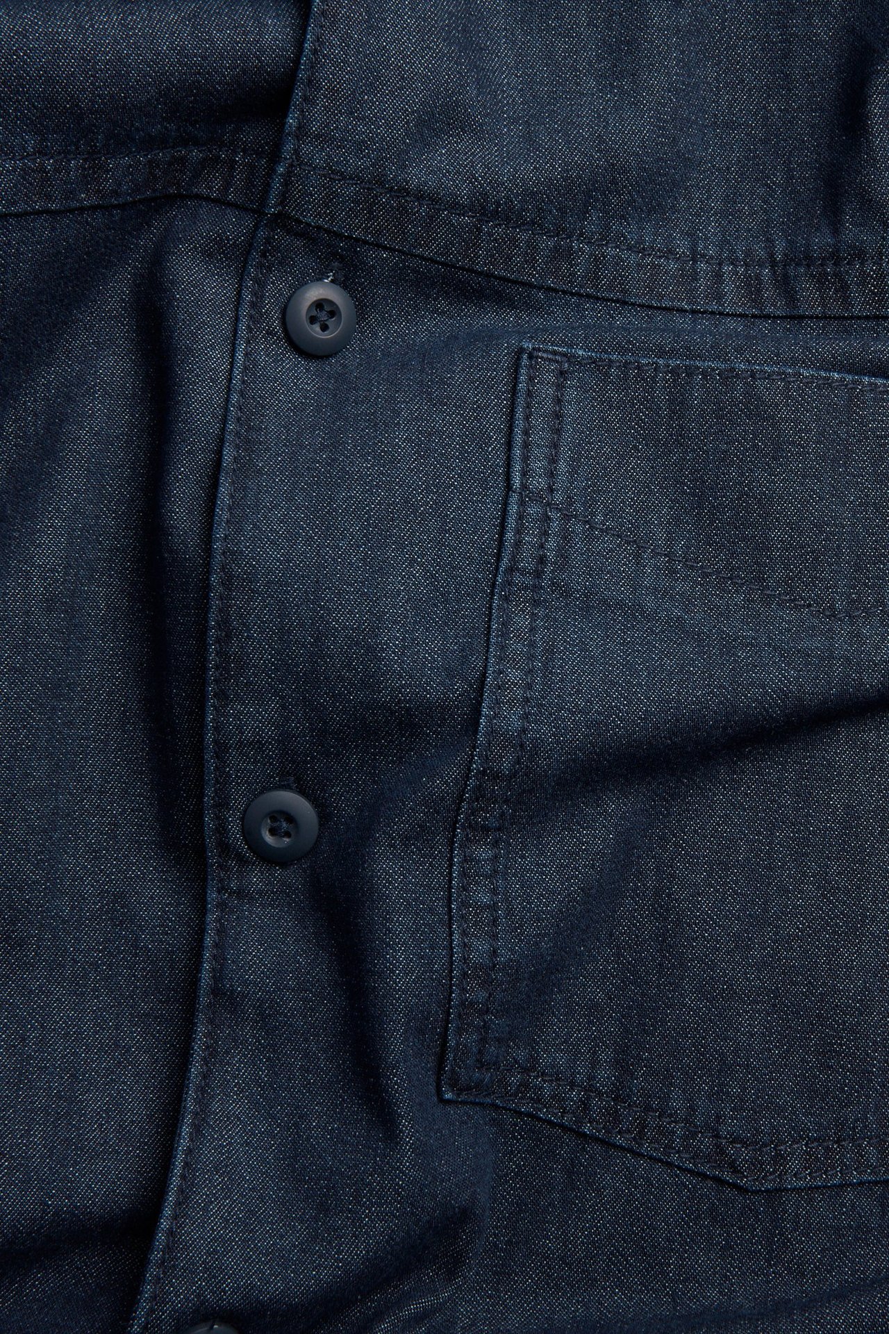 Närbild framifrån på en mörkblå overshirt i denim med knäppning och en bröstficka.