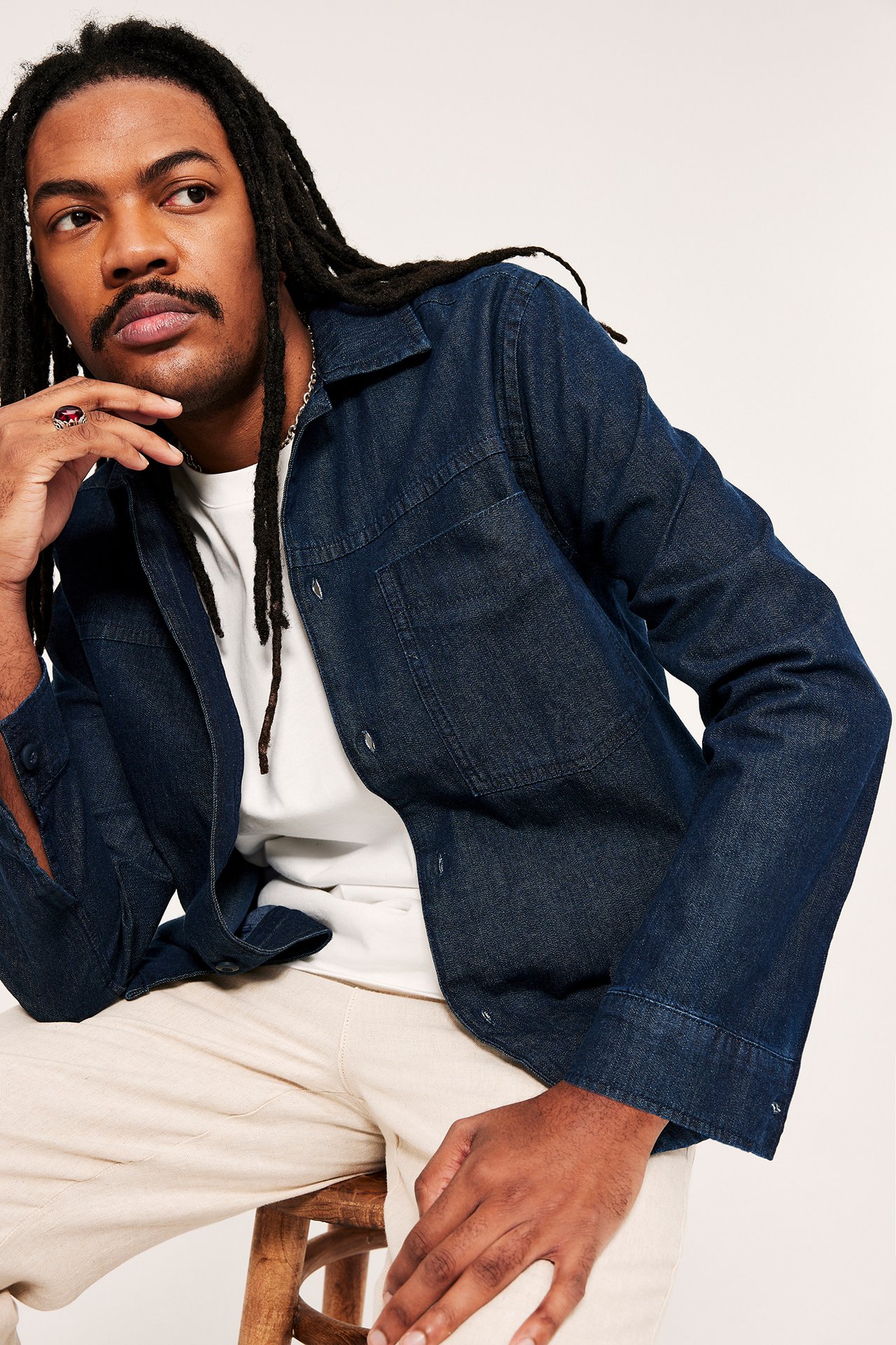 Lähikuva miehestä yllään avoin tummansininen farkkuovershirt valkoisen t-paidan ja vaaleanbeigien housujen päällä.