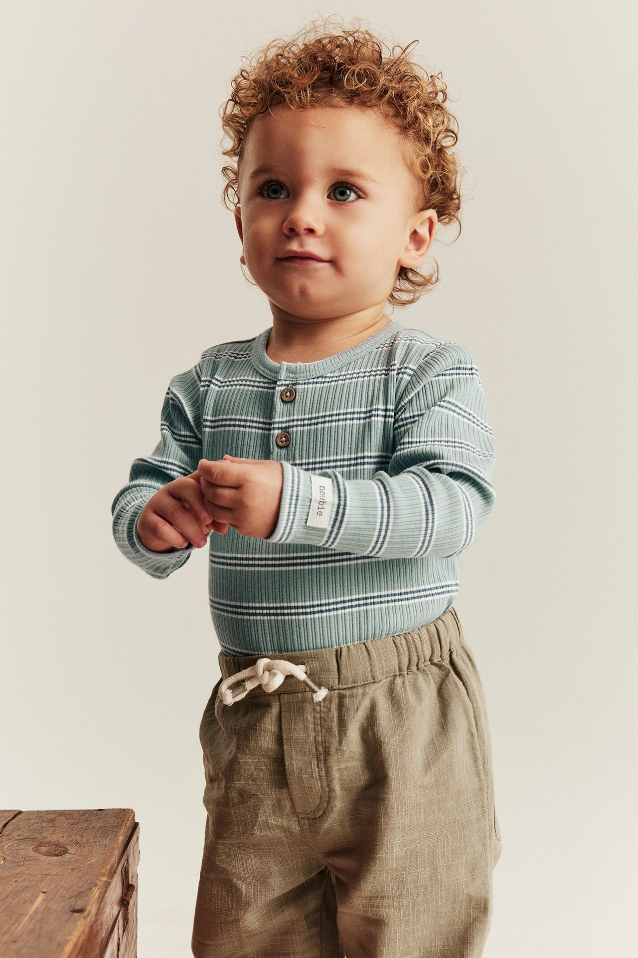 Framifrån: baby bär en ljusgrön randig ribbad body med knappar och ljusbruna byxor.