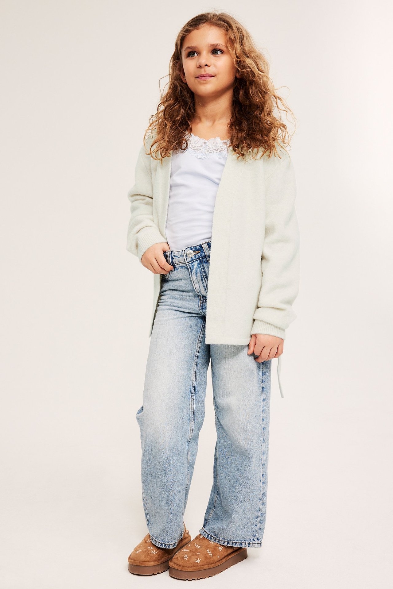 Edestä: Lapsi valkoisessa meleeratussa neuletakissa, valkoinen pitsitoppi, vaaleansiniset farkut, ruskeat tohvelit.