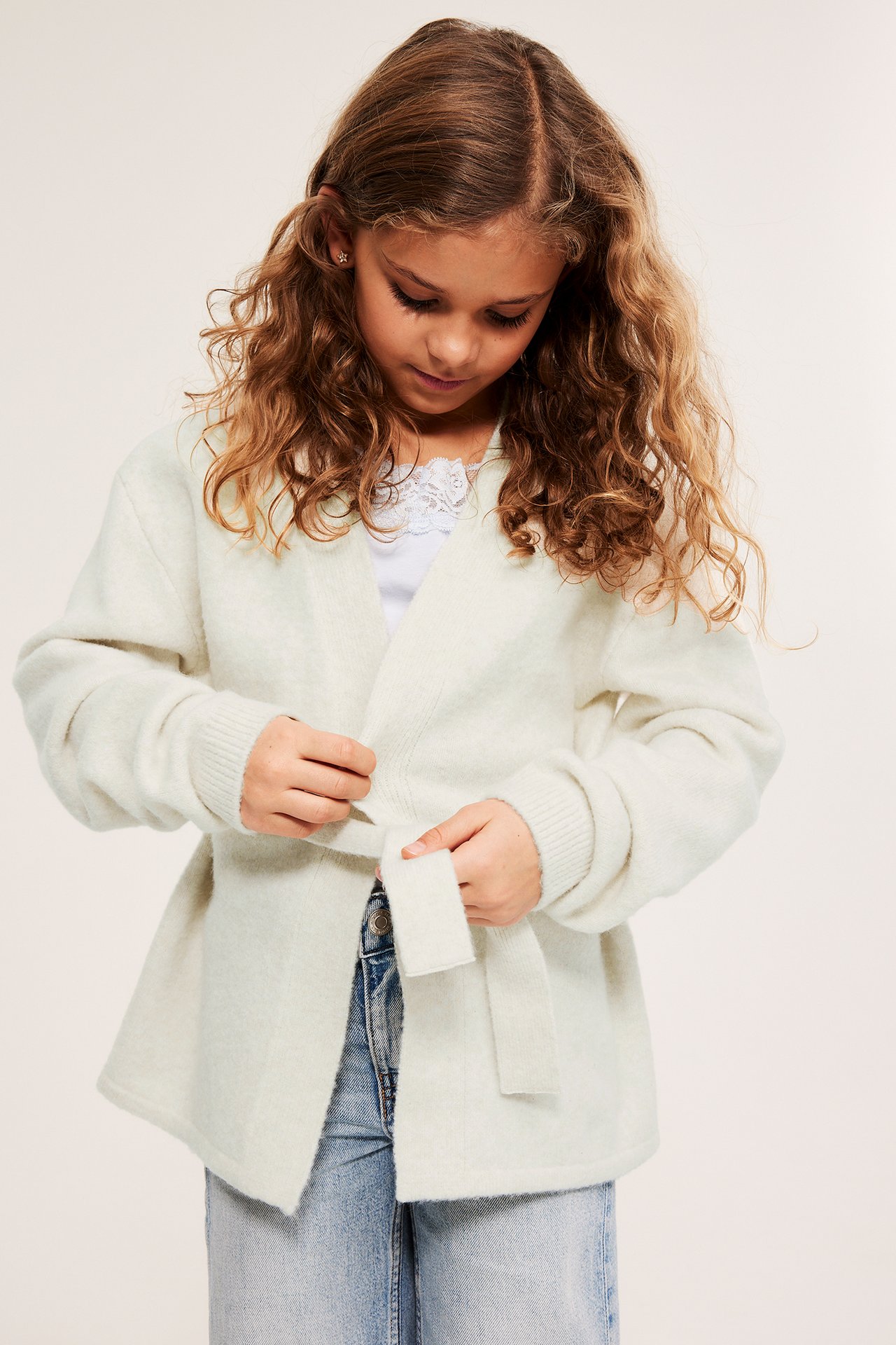 Edestä kuvattu lapsi sitomassa valkoista melange-neuletakkia valkoisen pitsitopin ja farkkujen yli.