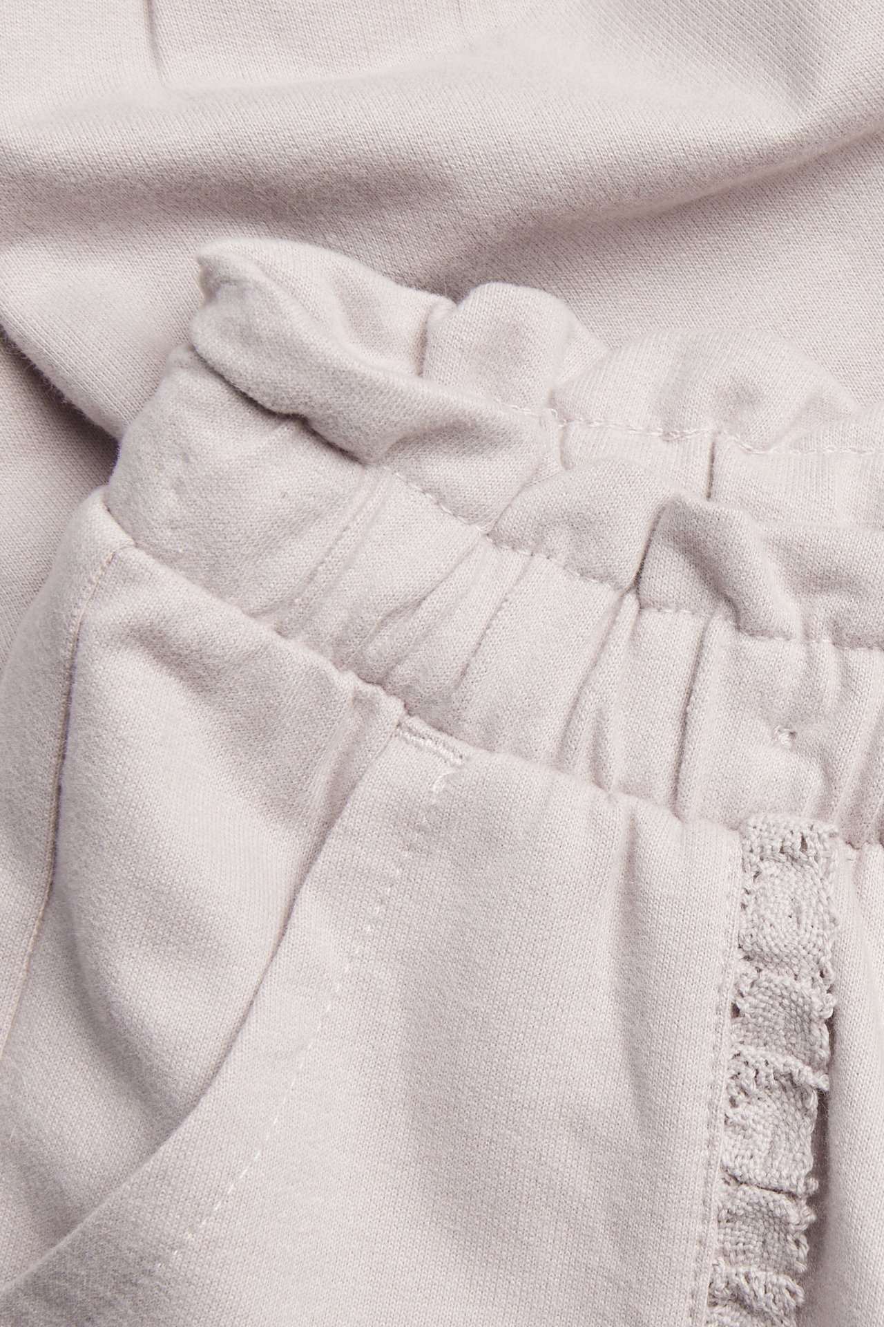 Nærbilde av lys lilla ensfargede joggebukser i bomull med elastisk midje og blondekant rundt lommene.
