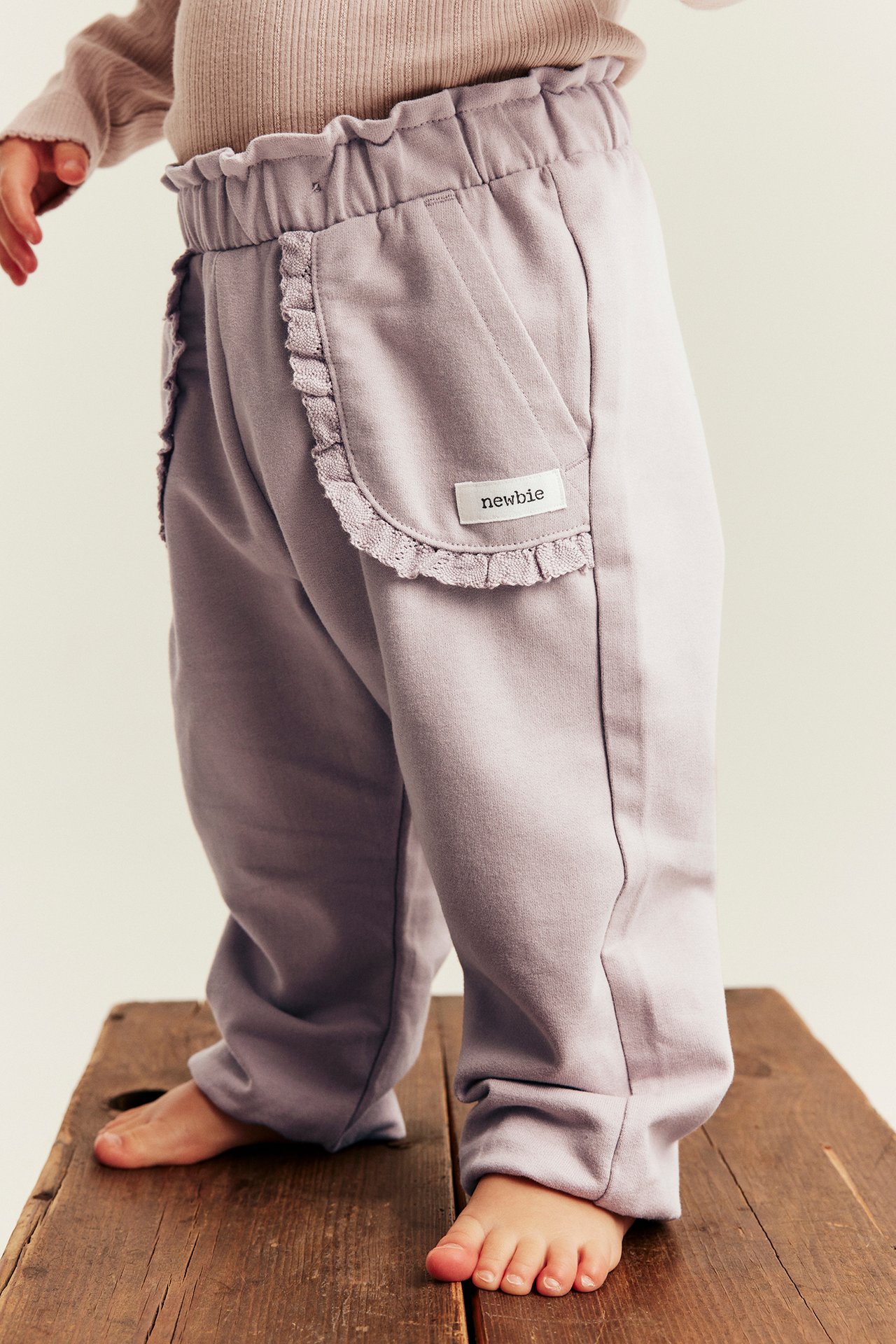 Sidebilde av babybukse i lys lilla med rysjemidje og volanglommer, sammen med en beige topp.