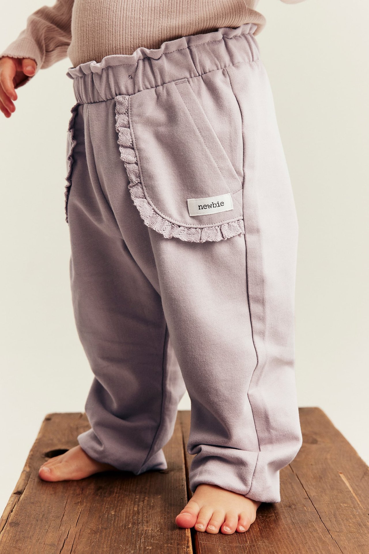Sidebilde av babybukse i lys lilla med rysjemidje og volanglommer, sammen med en beige topp.