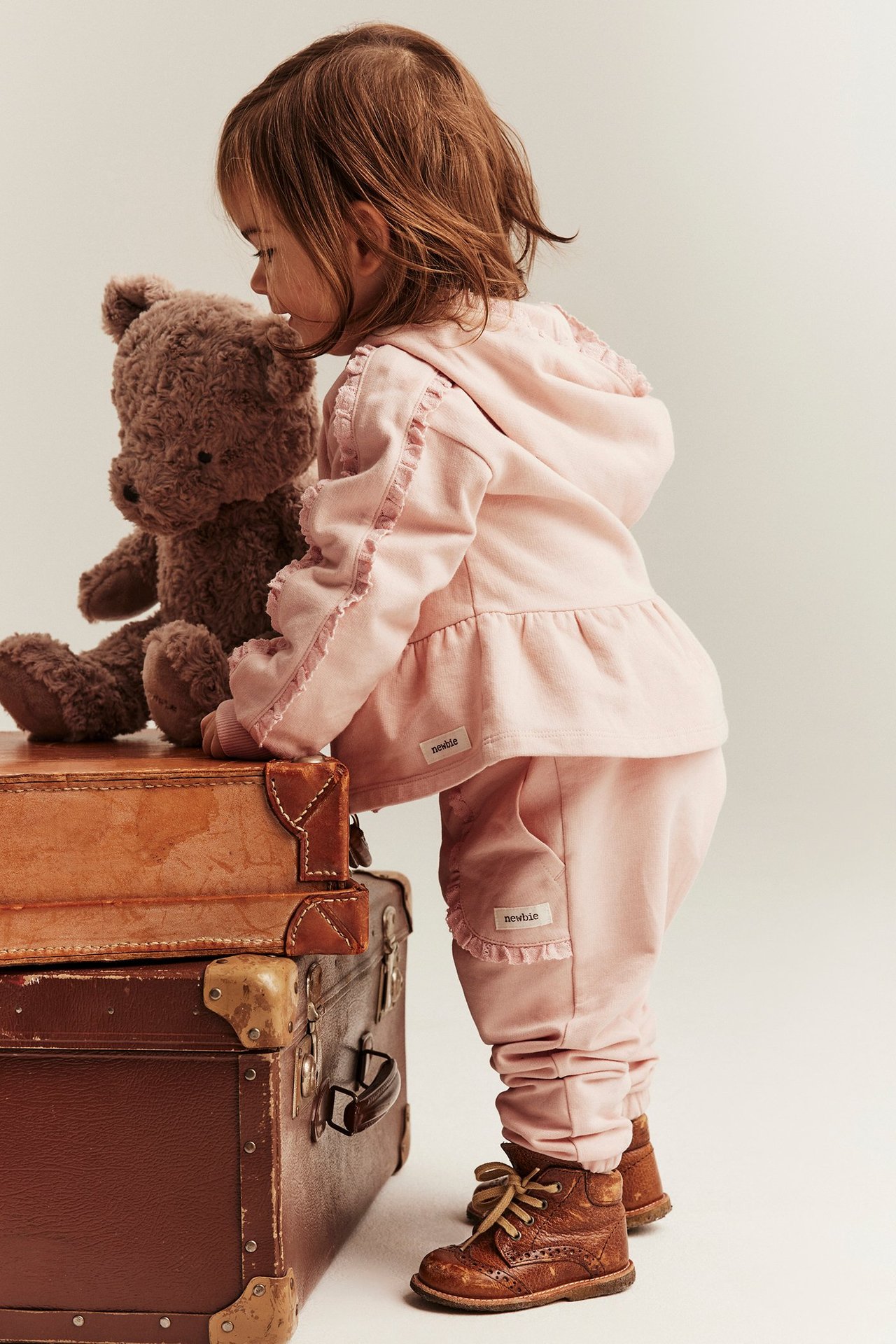 Sidevisning av et barn i lys rosa bomullsdress med volangdetaljer og brune snørestøvler.