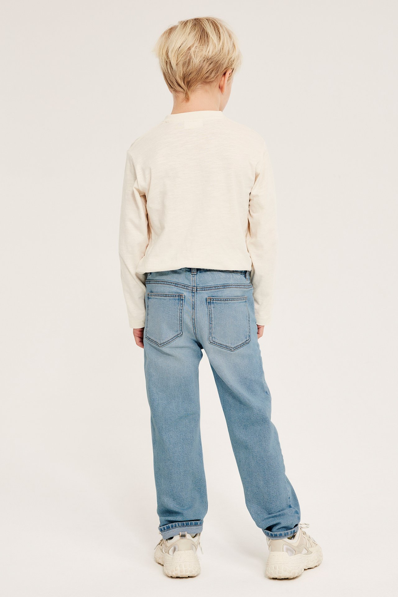 Widok z tyłu dziecka w jasnoniebieskich dżinsach relaxed fit, jasnokremowej koszulce i beżowych sneakersach.