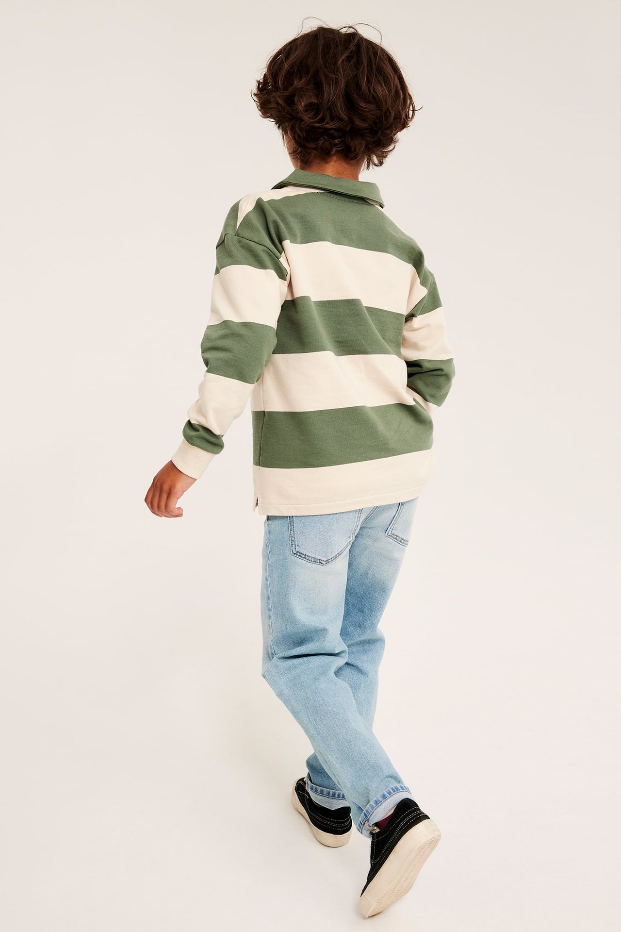 Ryggbilde av et barn i grønn- og beigestripet langermet pologenser, lyseblå jeans og svarte sneakers.