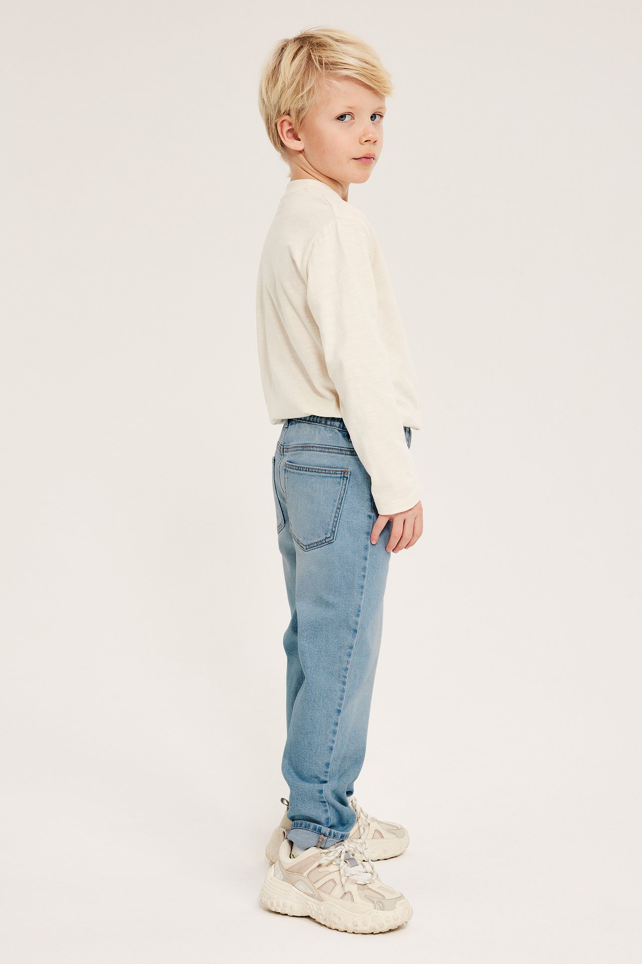 Sidovy av ett barn i ljusblå relaxed fit denimjeans, en gräddvit långärmad tröja och beiga sneakers.