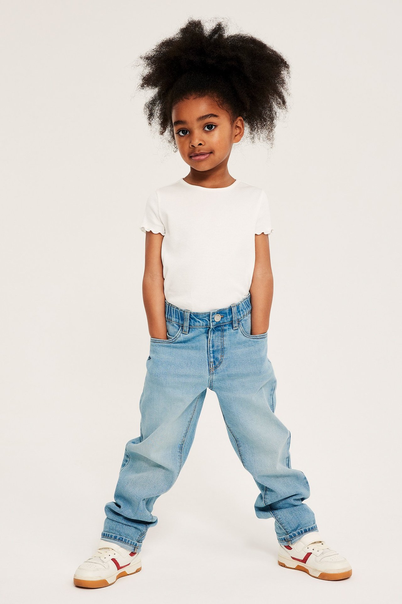 Widok z przodu dziecka w jasnoniebieskich luźnych jeansach, białym prążkowanym t-shircie z falbankami i białych sneakersach.