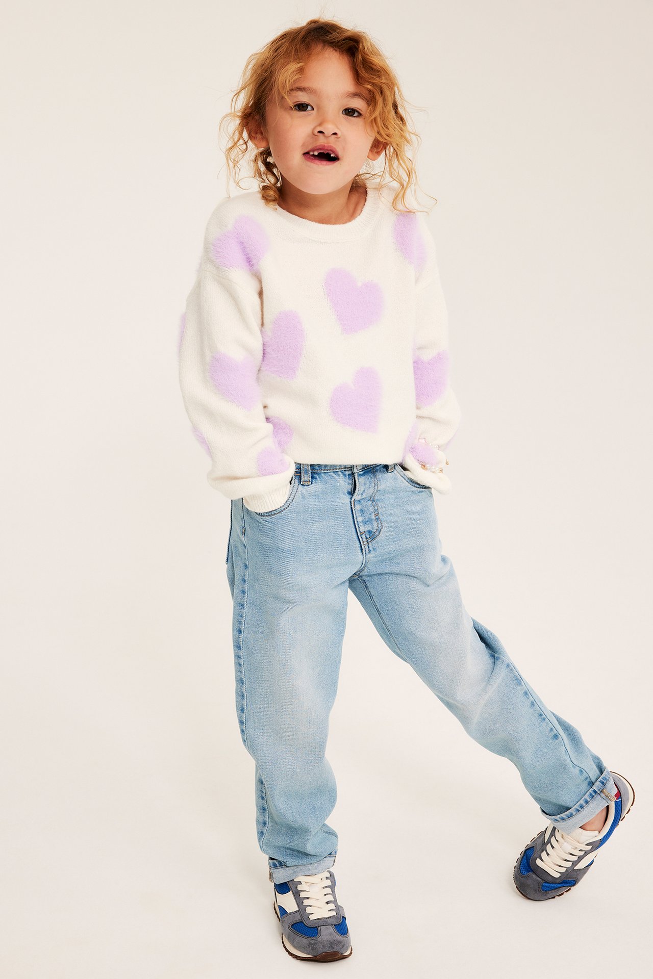 Framifrån barn bär en vit luddig tröja med lila hjärtan, ljusblå jeans och blåa sneakers.