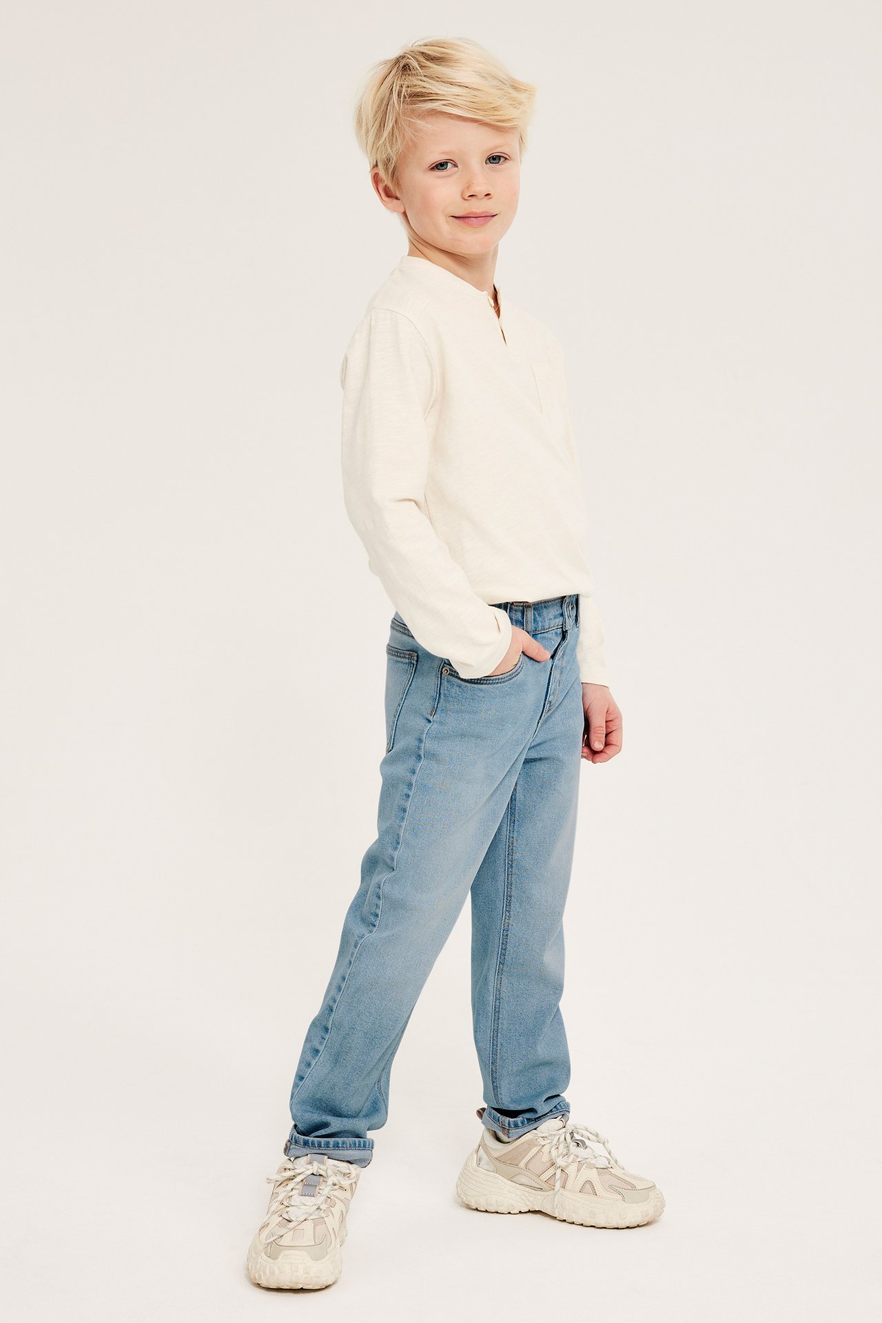 Widok z boku: Dziecko w jasnoniebieskich jeansach relaxed, jasnobeżowej koszulce z długim rękawem i beżowych sneakersach.