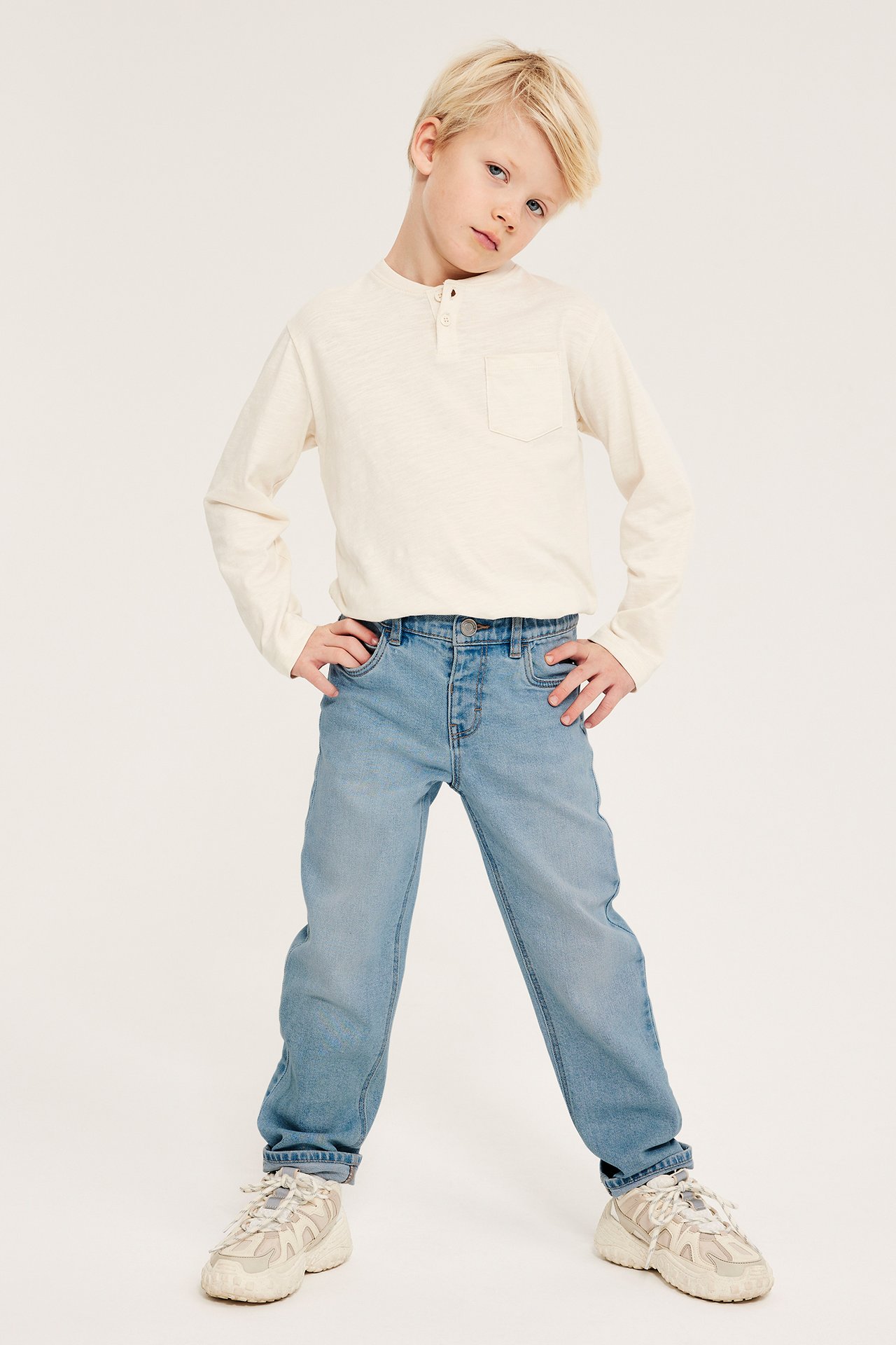 Frontvy av ett barn i ljusblå relaxed fit denimjeans, krämvit långärmad tröja och beiga sneakers.