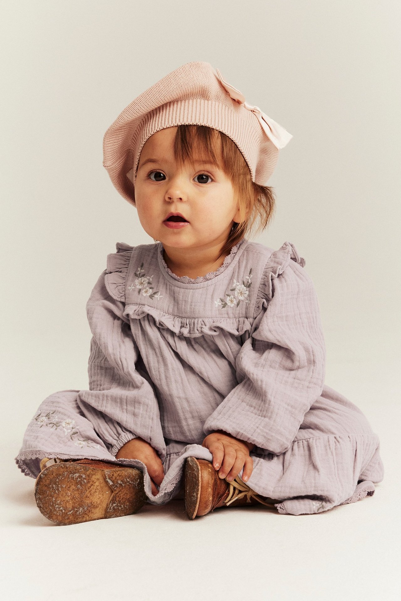 Framsida: Baby bär en ljuslila vävd klänning med broderade blommor, volanger, puffärmar och rosa basker.
