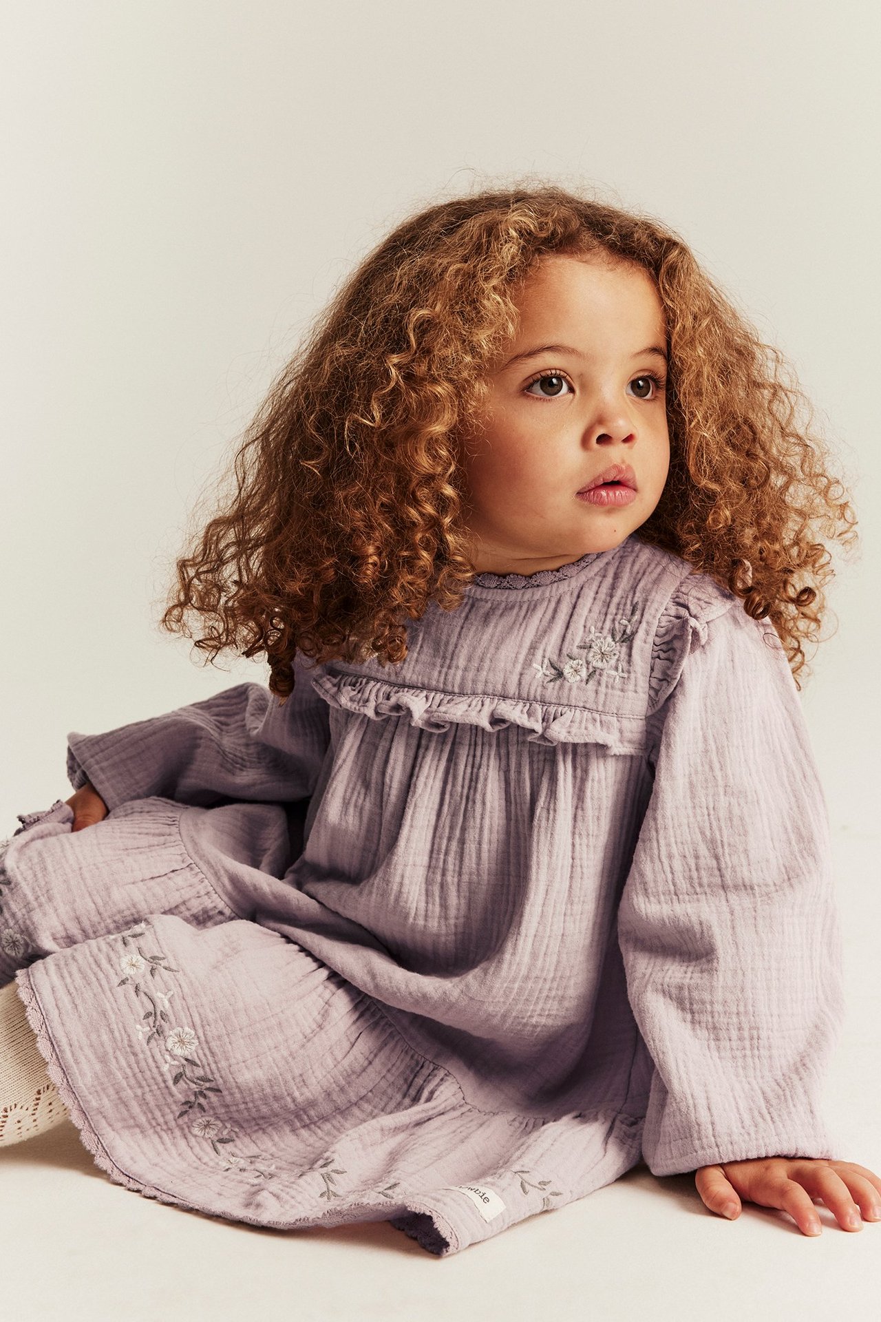 Widok z przodu: niemowlę w jasnoliliowej bawełnianej sukience z falbanką, haftem kwiatowym i białymi skarpetkami.