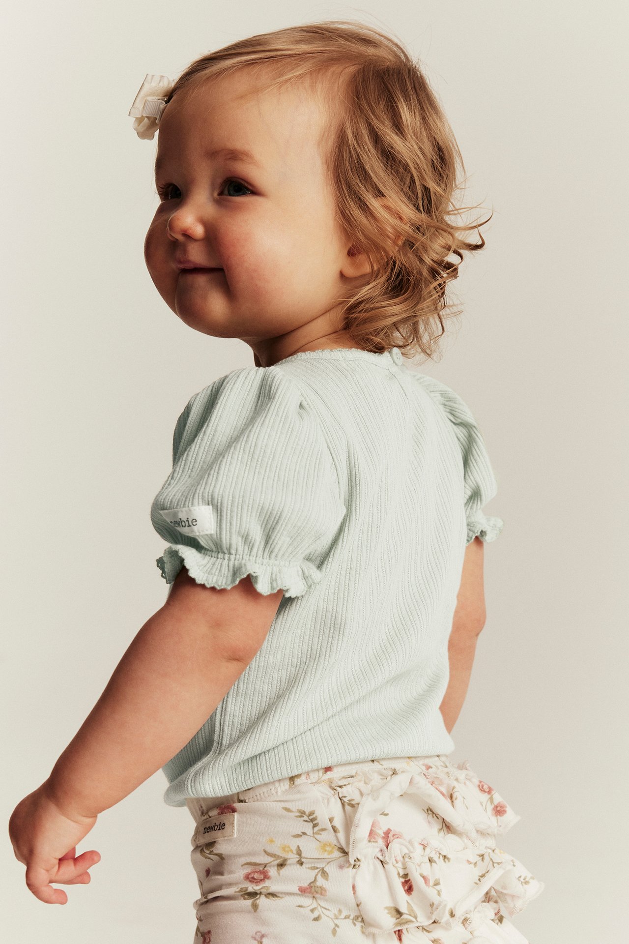 Sidovy av baby i ljusgrön ribbad body med puffärm och vita blommiga volangtrosor med rosett i håret.
