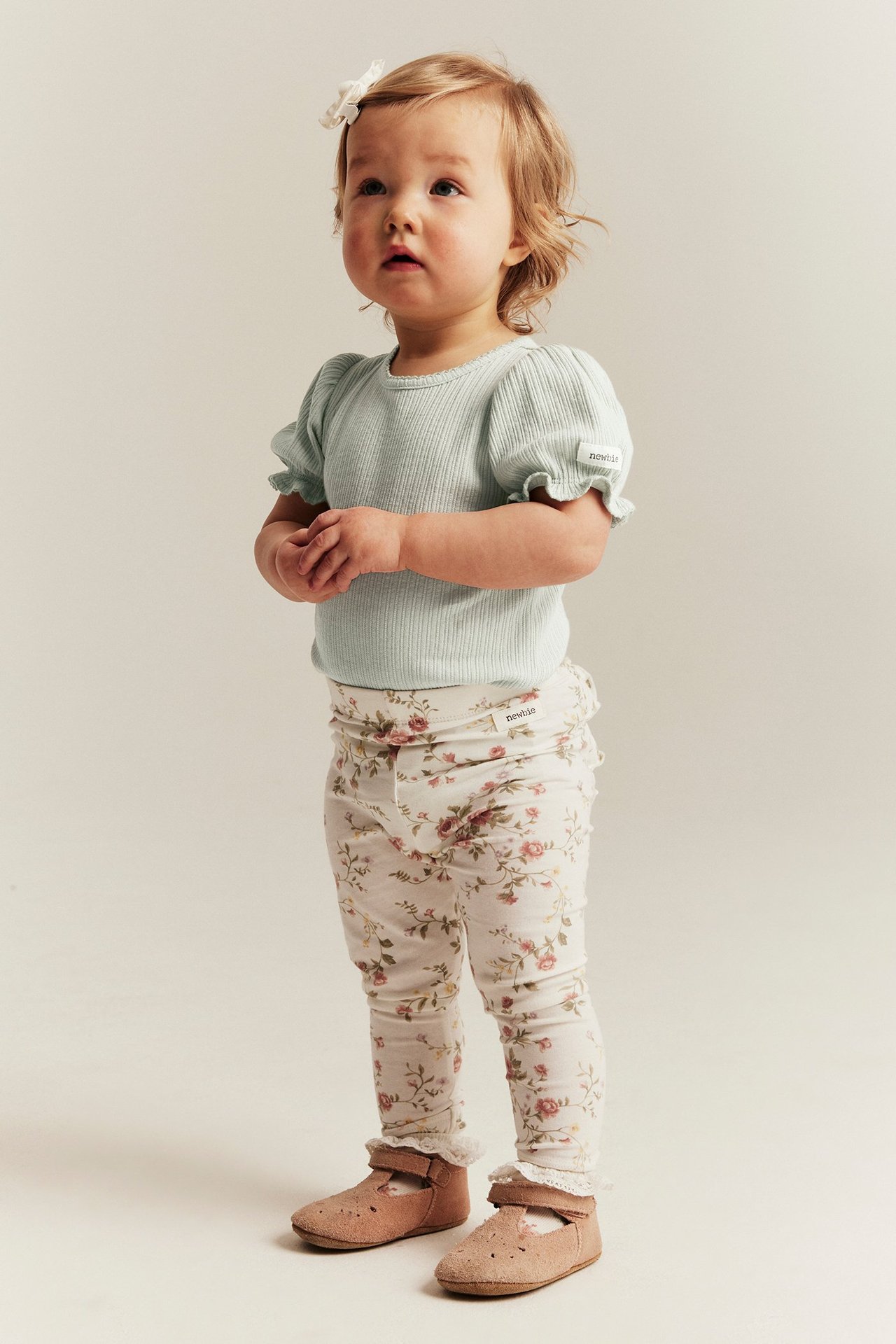 Front view av en baby i ljusgrön ribbad puffärmsbody, krämvita blommiga leggings och rosa skor.