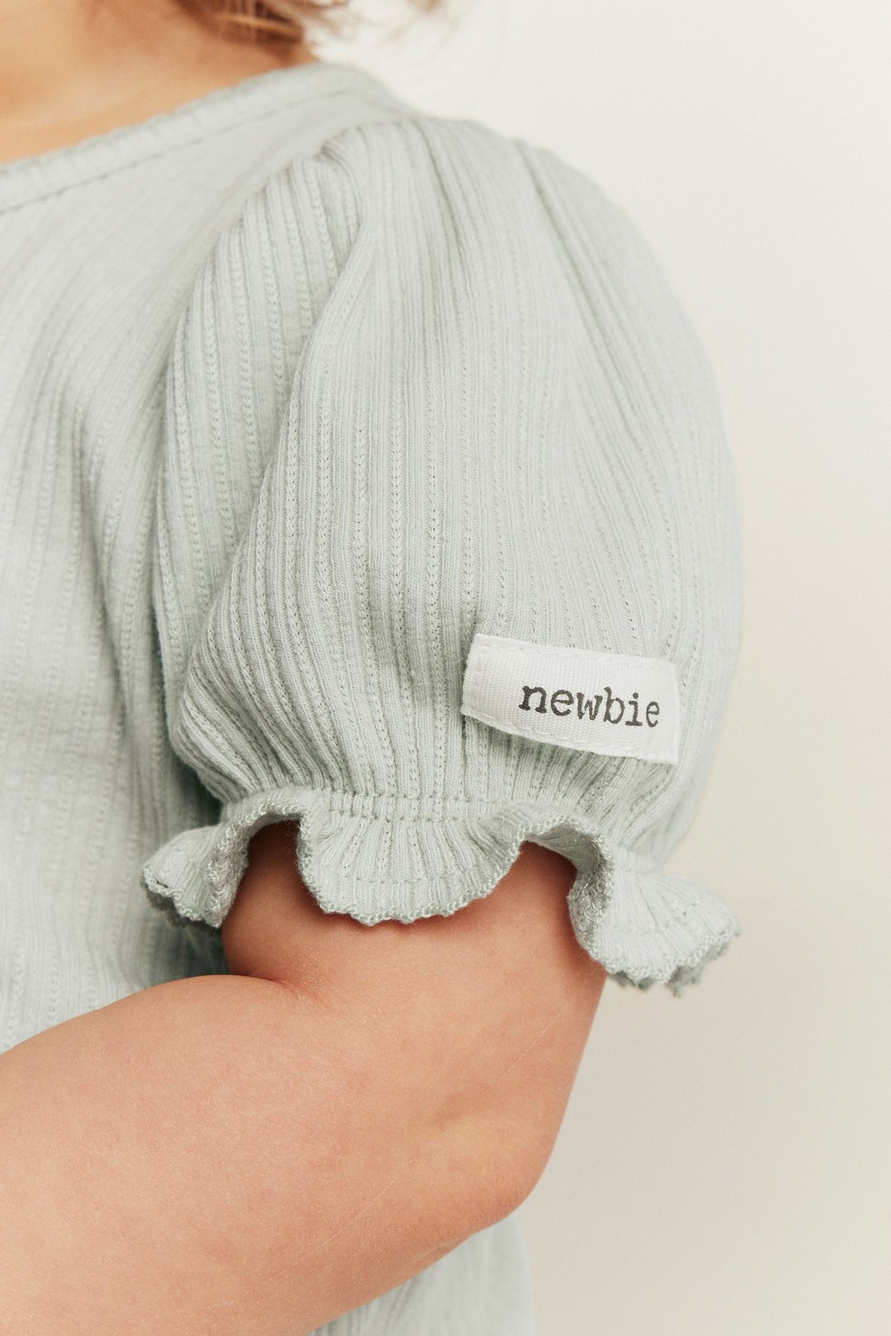Närbild: Baby i en ljusgrön ribbad jerseybody med puffärm och volangkanter. Newbie-etikett.
