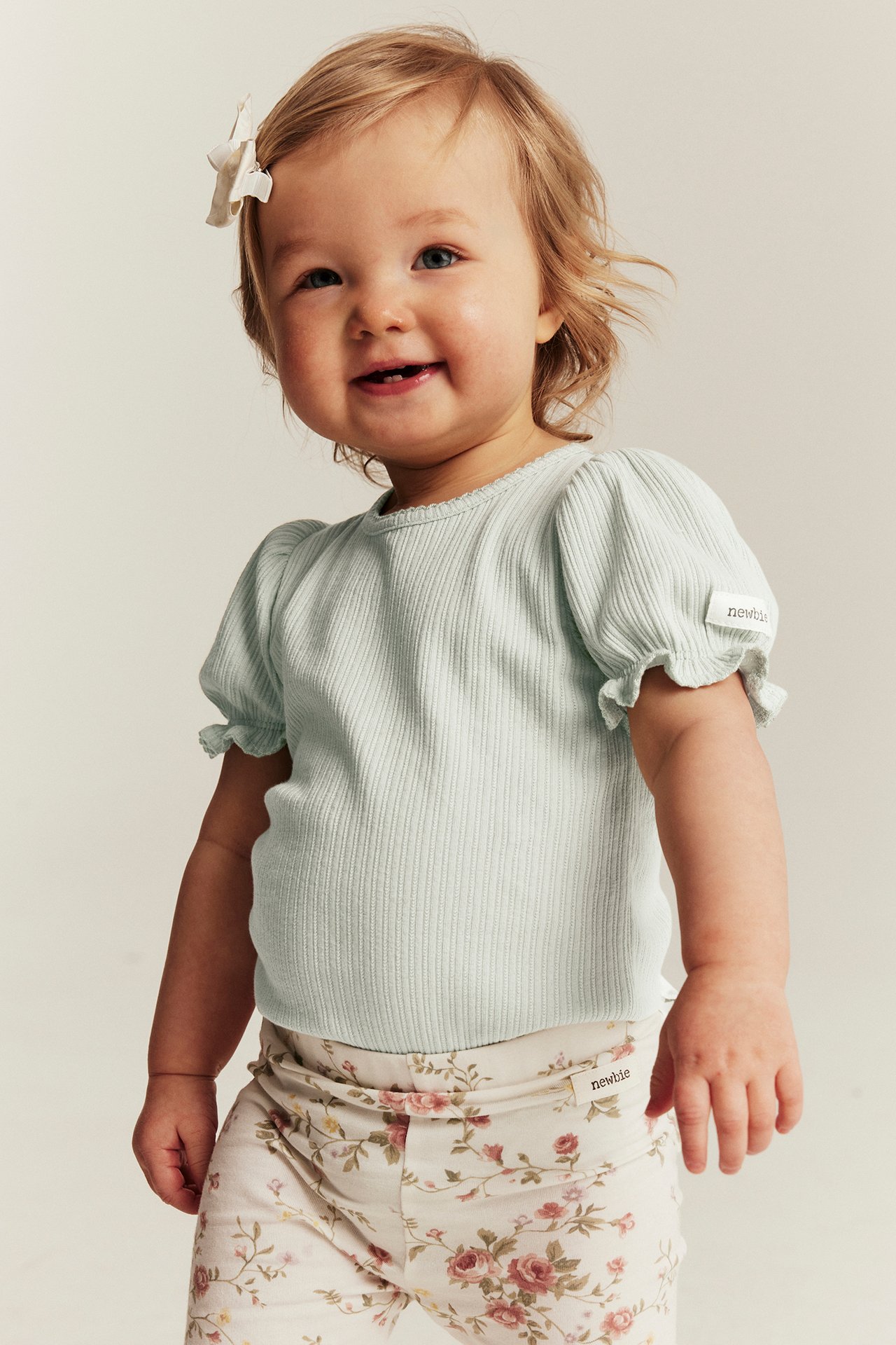 Frontvy av baby i en ljusgrön ribbad body med puffärm och benvita blommiga byxor.