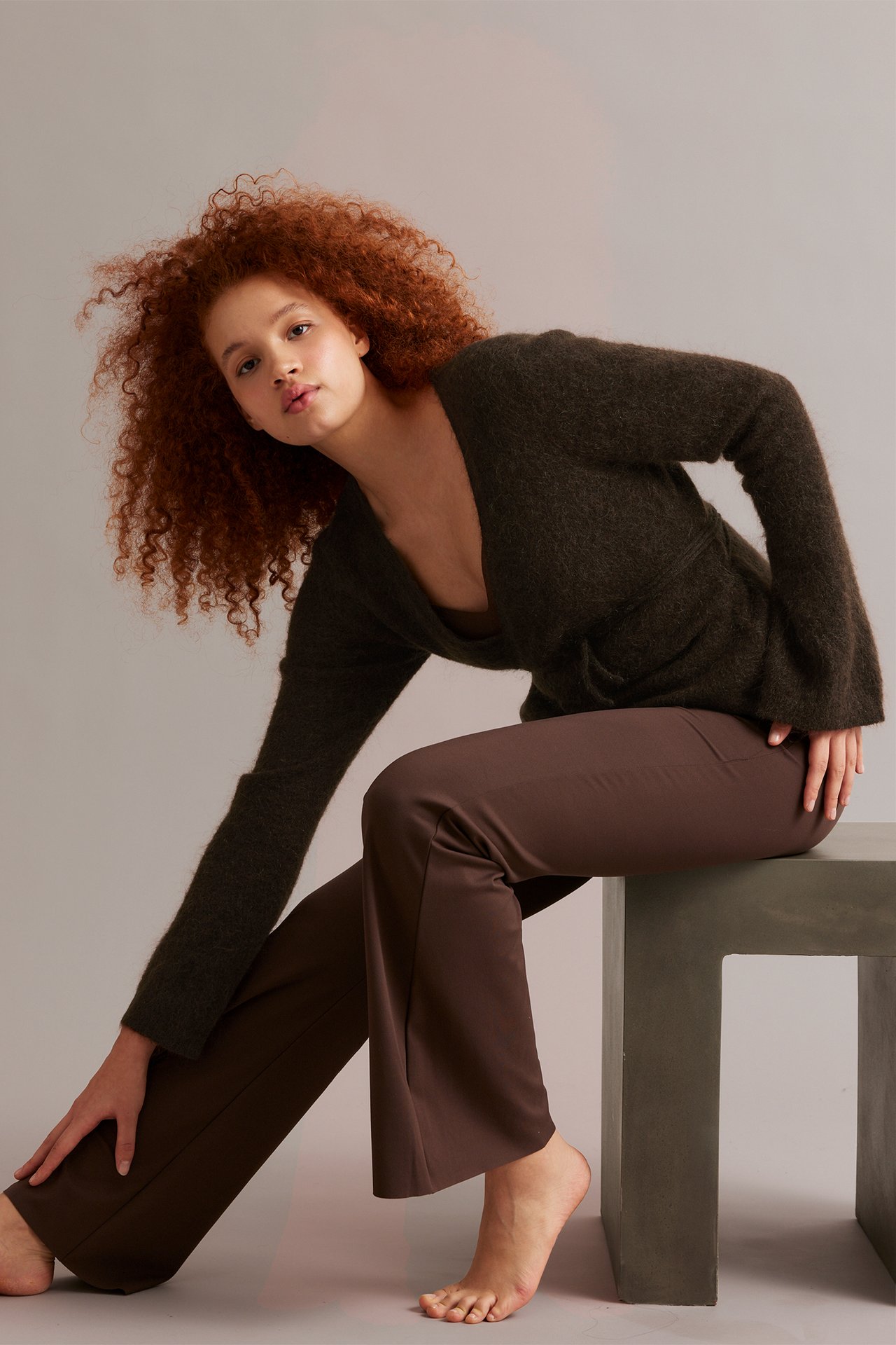 Widok z przodu kobiety w brązowym wełnianym kardiganie kopertowym z szerokimi rękawami i brązowych spodniach.