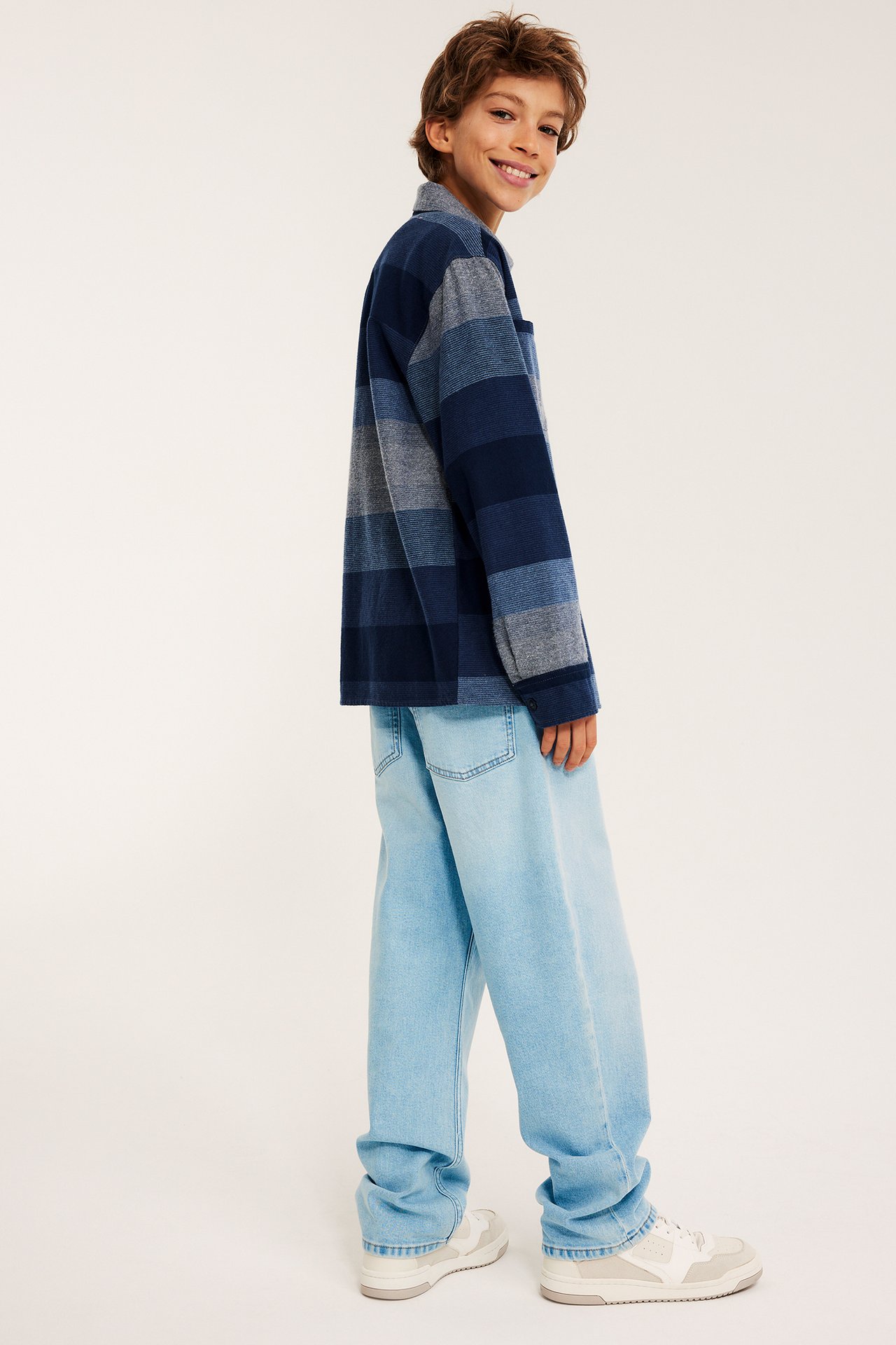 Widok z boku dziecka w jasnoniebieskich luźnych jeansach, pasiastej koszuli, białych trampkach.