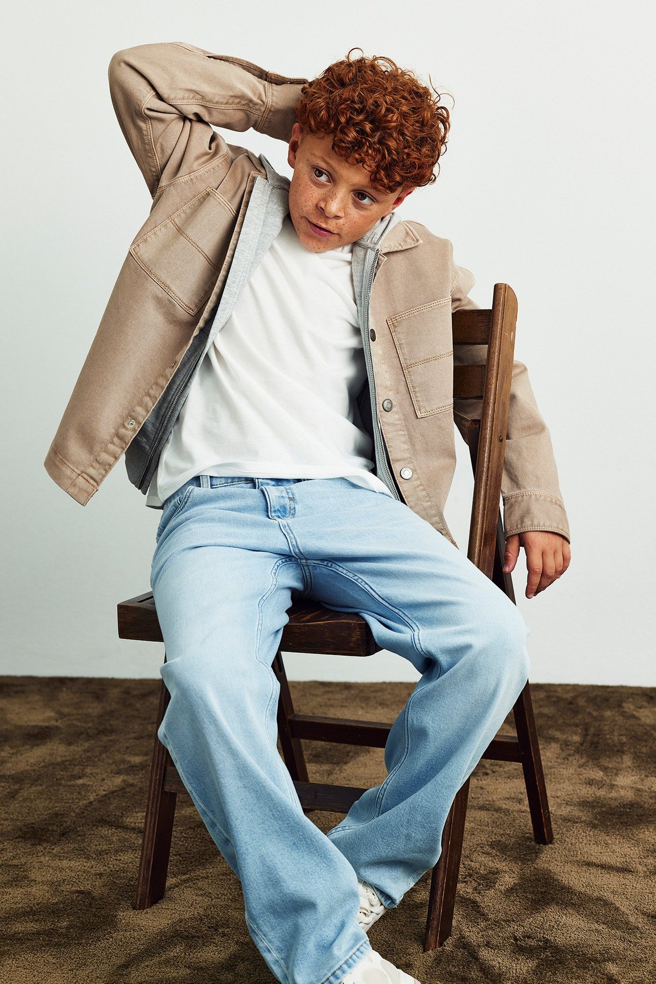 Edestä: Lapsi vaaleansinisissä löysissä farkuissa, valkoinen t-paita, beige takki ja harmaa huppu.