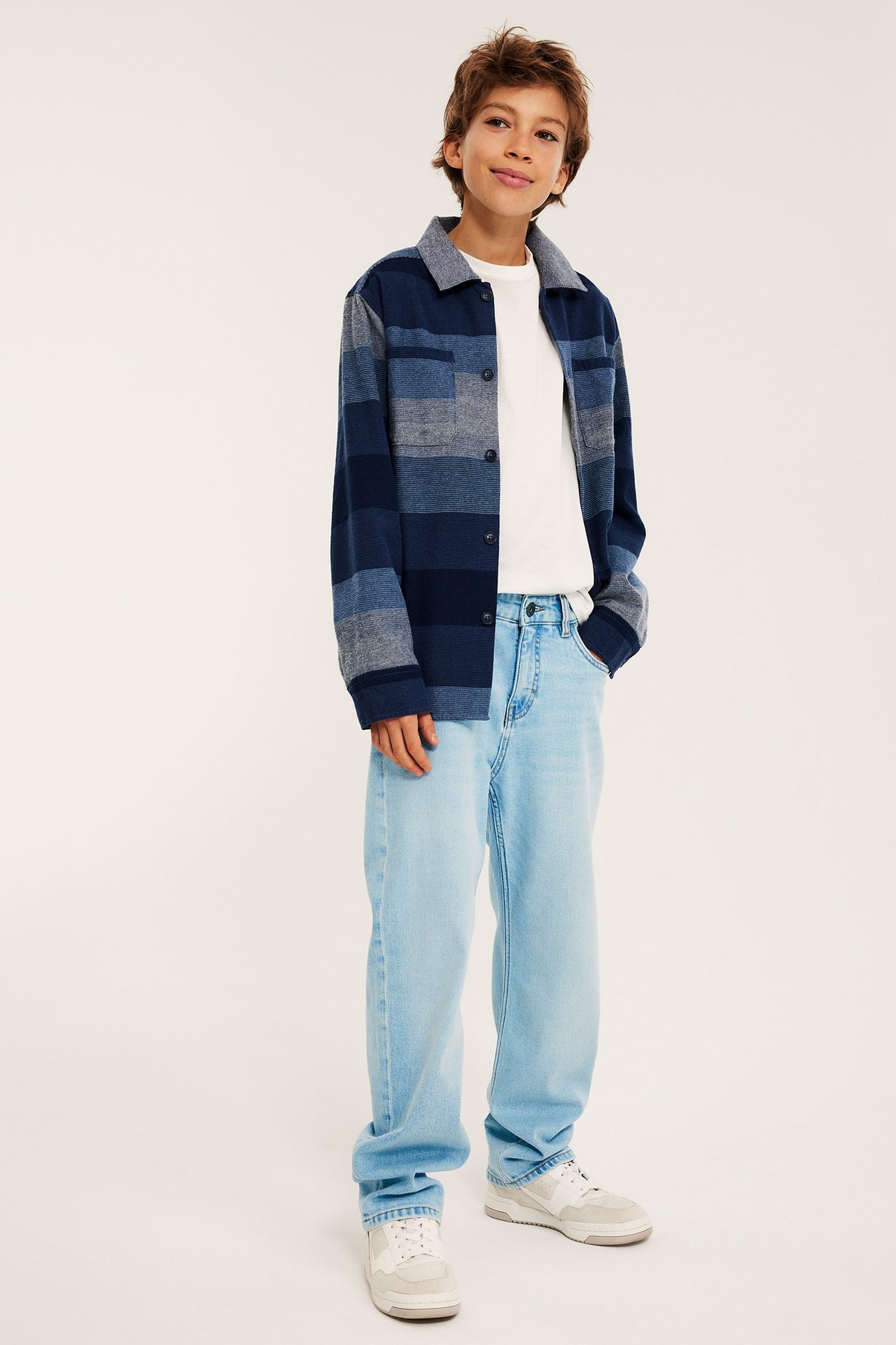 Widok z przodu: Chłopiec w jasnoniebieskich luźnych jeansach, pasiastej niebieskiej koszuli-kurtce, białym T-shircie i trampkach.