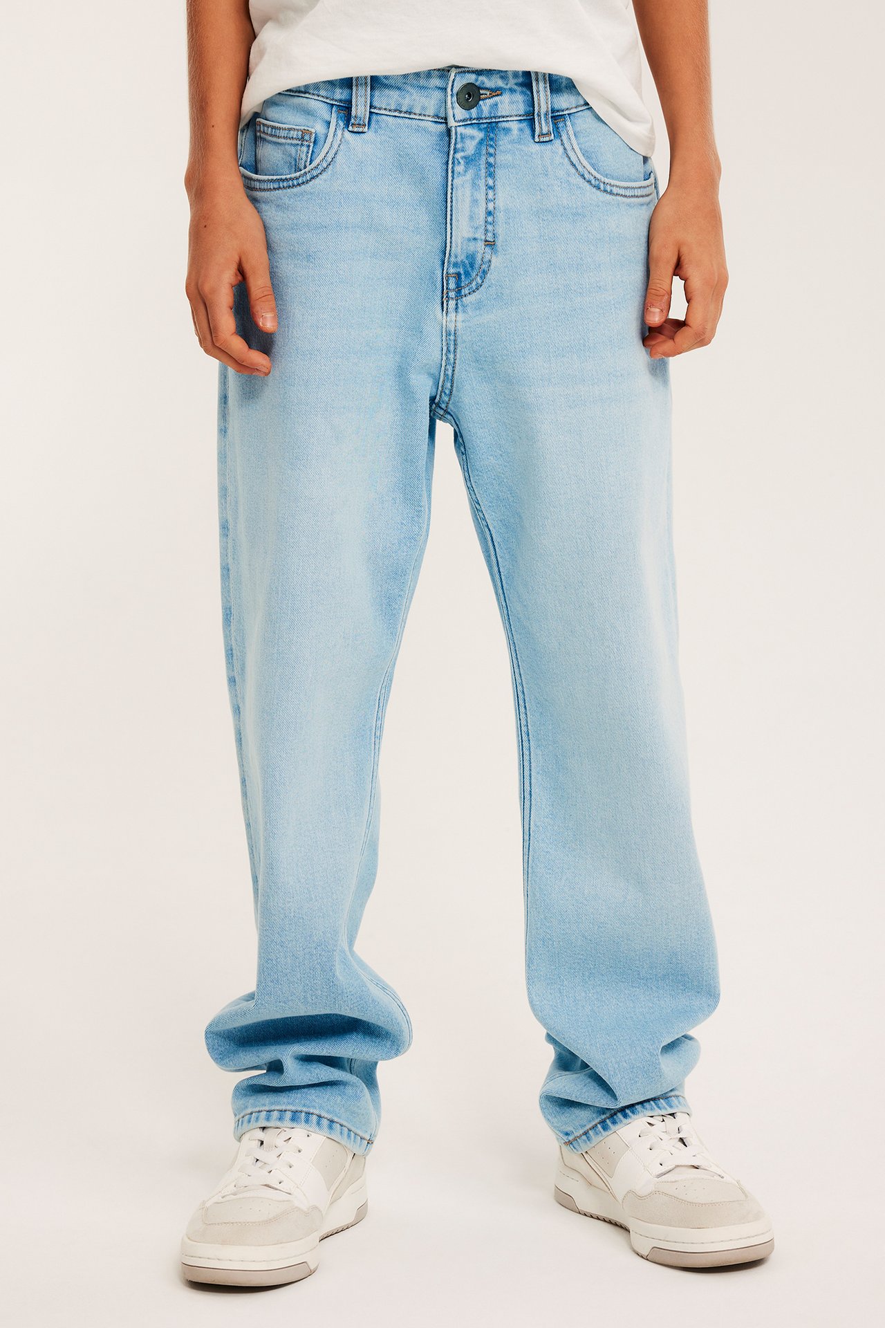 Widok z przodu dziecka w jasnych luźnych dżinsach, białym t-shircie i białobeżowych trampkach.