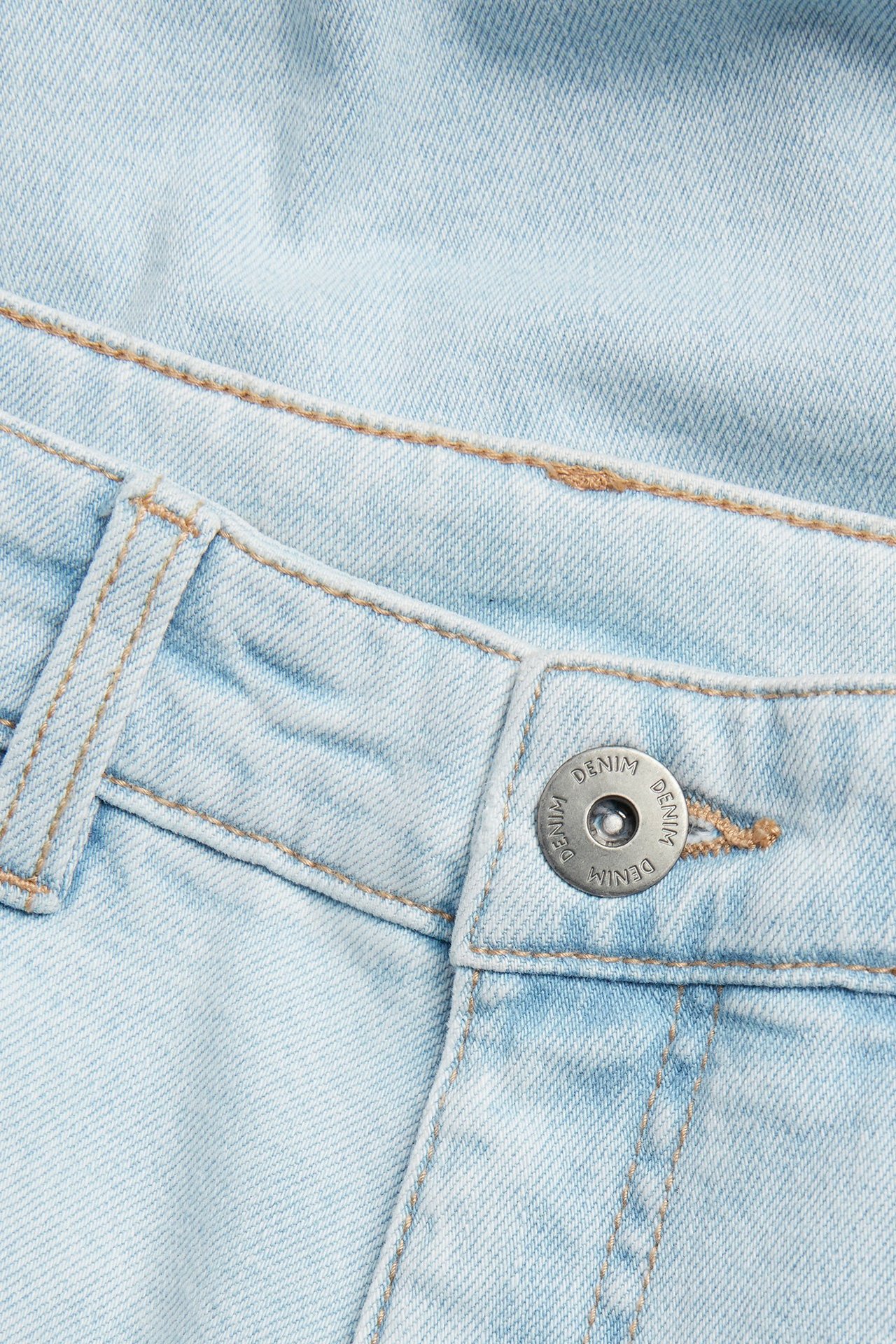 Zbliżenie na jasnoniebieskie luźne jeansy ze srebrnym guzikiem i jasnobrązowymi przeszyciami.
