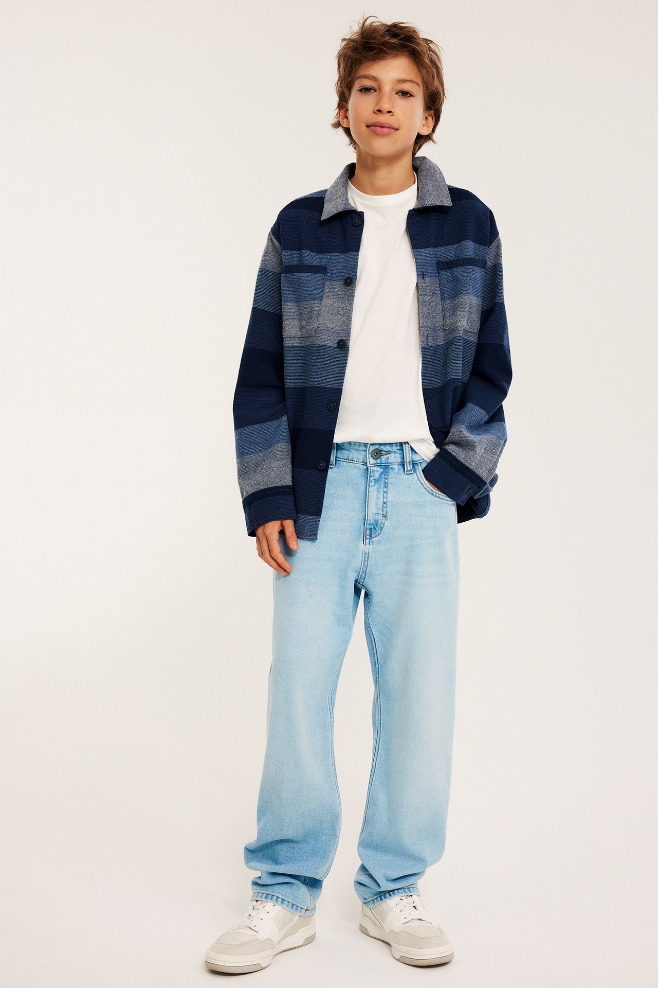 Widok z przodu dziecka w jasnoniebieskich luźnych jeansach, białym T-shircie i niebieskiej koszuli w paski.