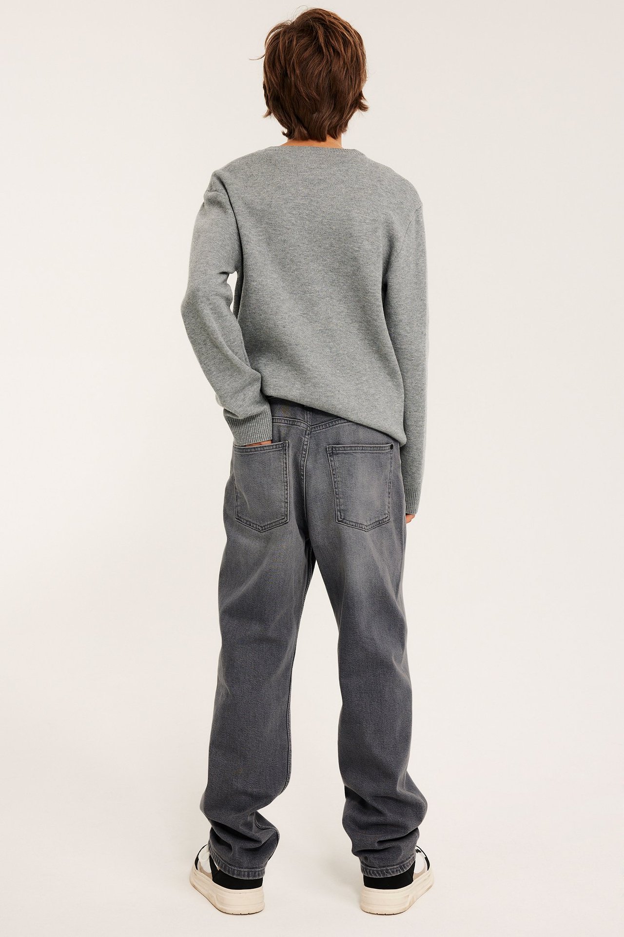 Widok z tyłu dziecka ubranego w jasnoszare luźne jeansy z regularnym stanem, szarą bluzę i czarno-białe sneakersy.