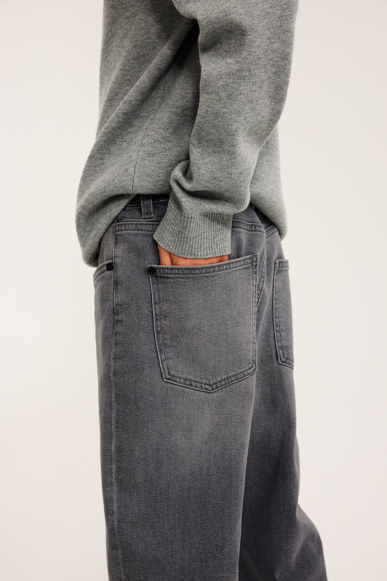 Widok z tyłu: Dziecko w jasnoszarych luźnych dżinsach, ręka w kieszeni, szary sweter.
