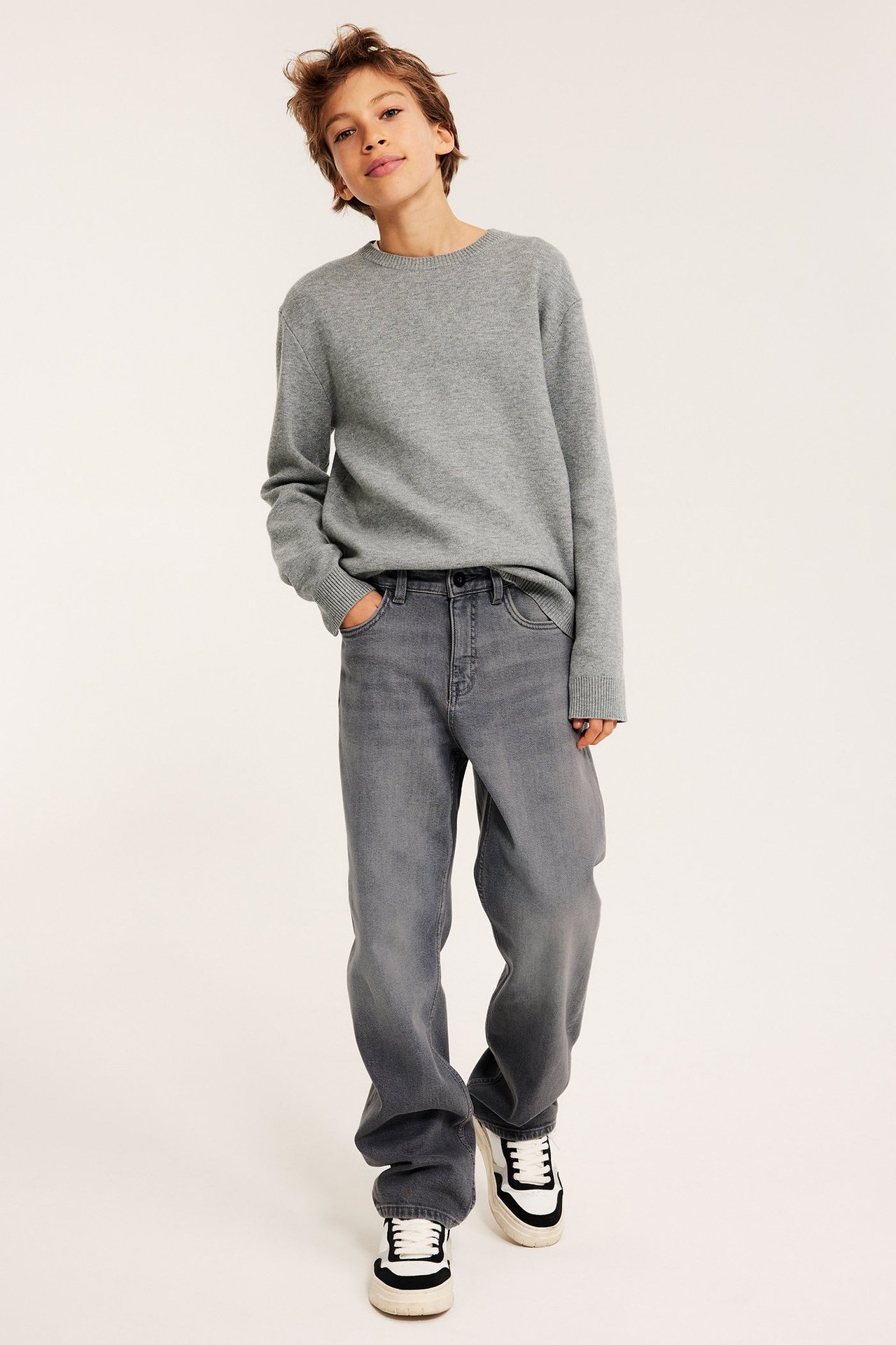 Widok z przodu dziecka w jasnoszarych luźnych jeansach z ręką w kieszeni, szarym swetrze i trampkach.