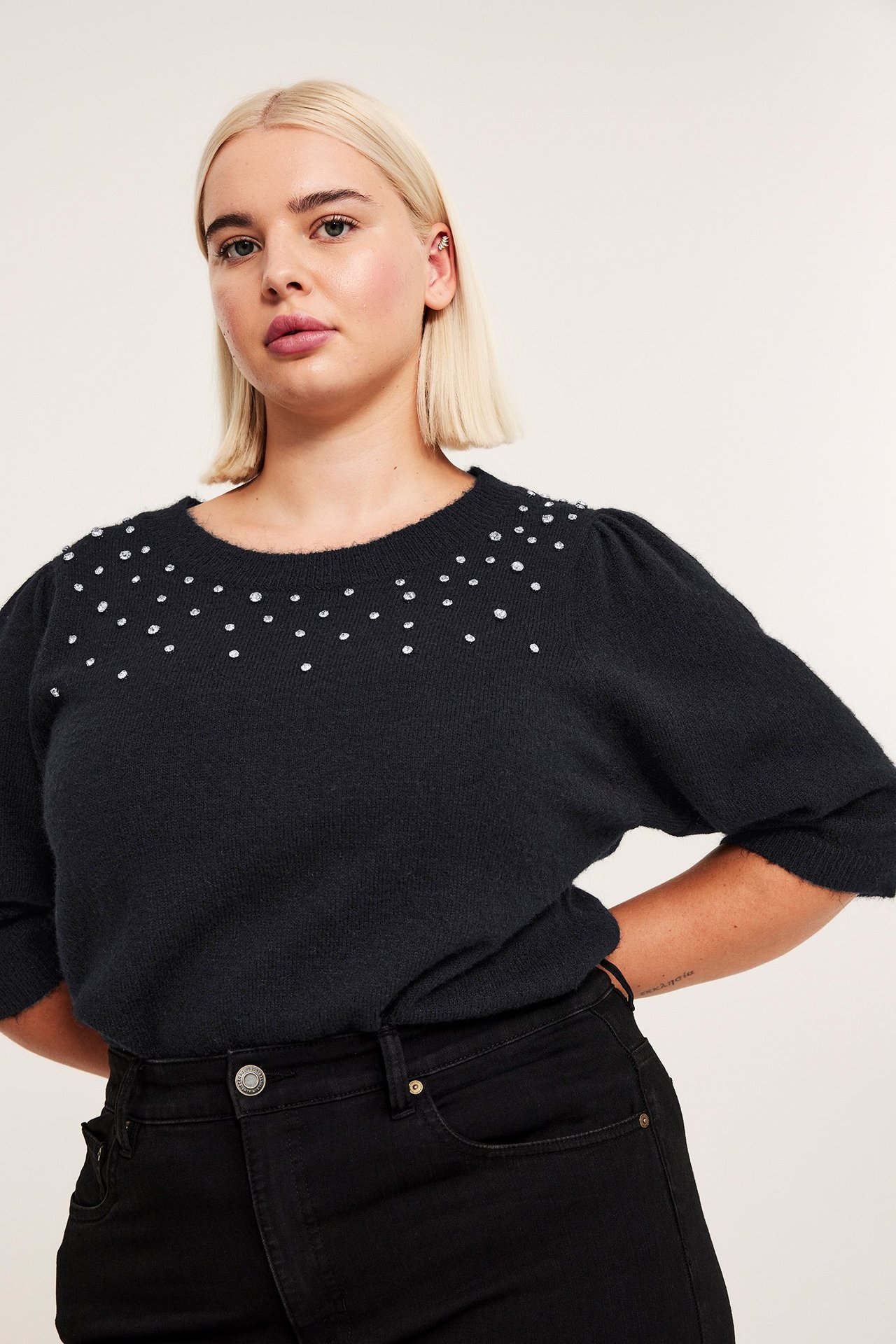 Widok z przodu: kobieta w czarnym swetrze z bufiastymi rękawami, cyrkoniami i czarnych dżinsach.