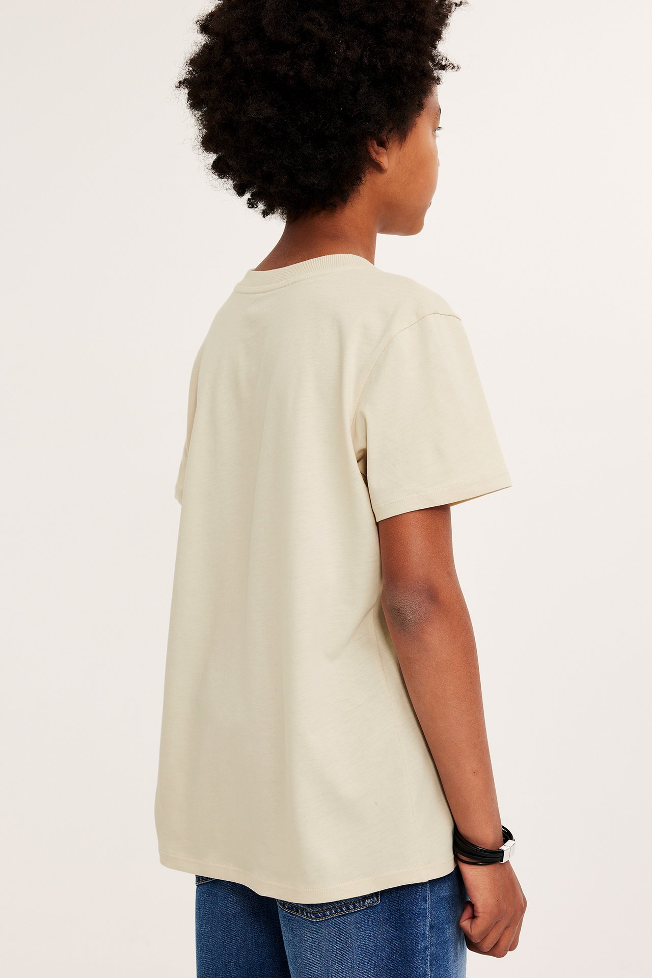 Sidovy av ett barn i en lös beige bomulls-t-shirt, blå jeans och ett svart armband.