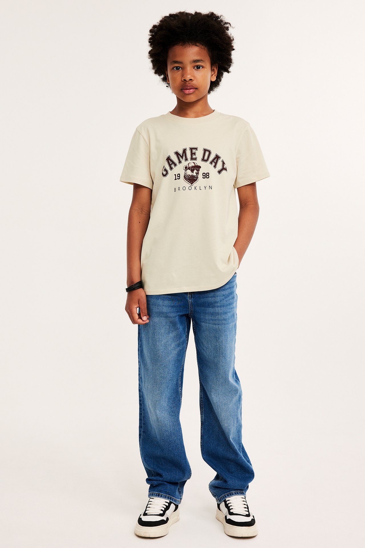 Framifrån: ett barn i beige bomulls-t-shirt med "Game Day"-tryck, blå jeans och sneakers.