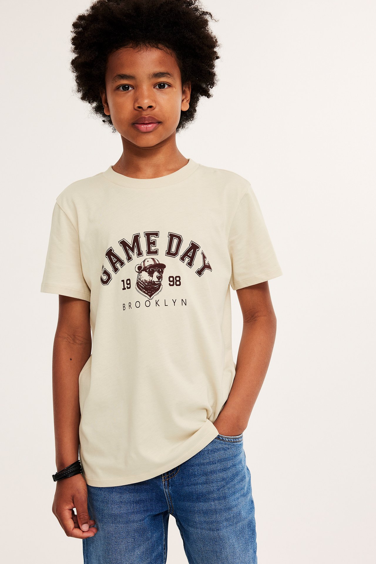 Frontvy: Ett barn i en beige bomulls-T-shirt med "GAME DAY"-tryck, blå jeans och ett svart armband.