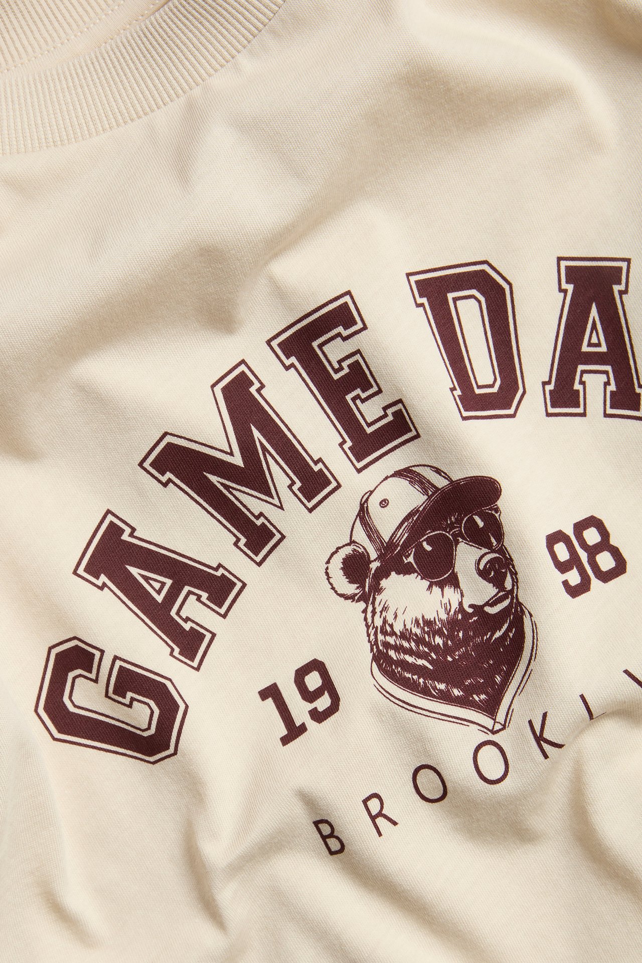 Närbild av en beige bomulls-t-shirt med brunt 'GAME DAY' och björn-tryck.