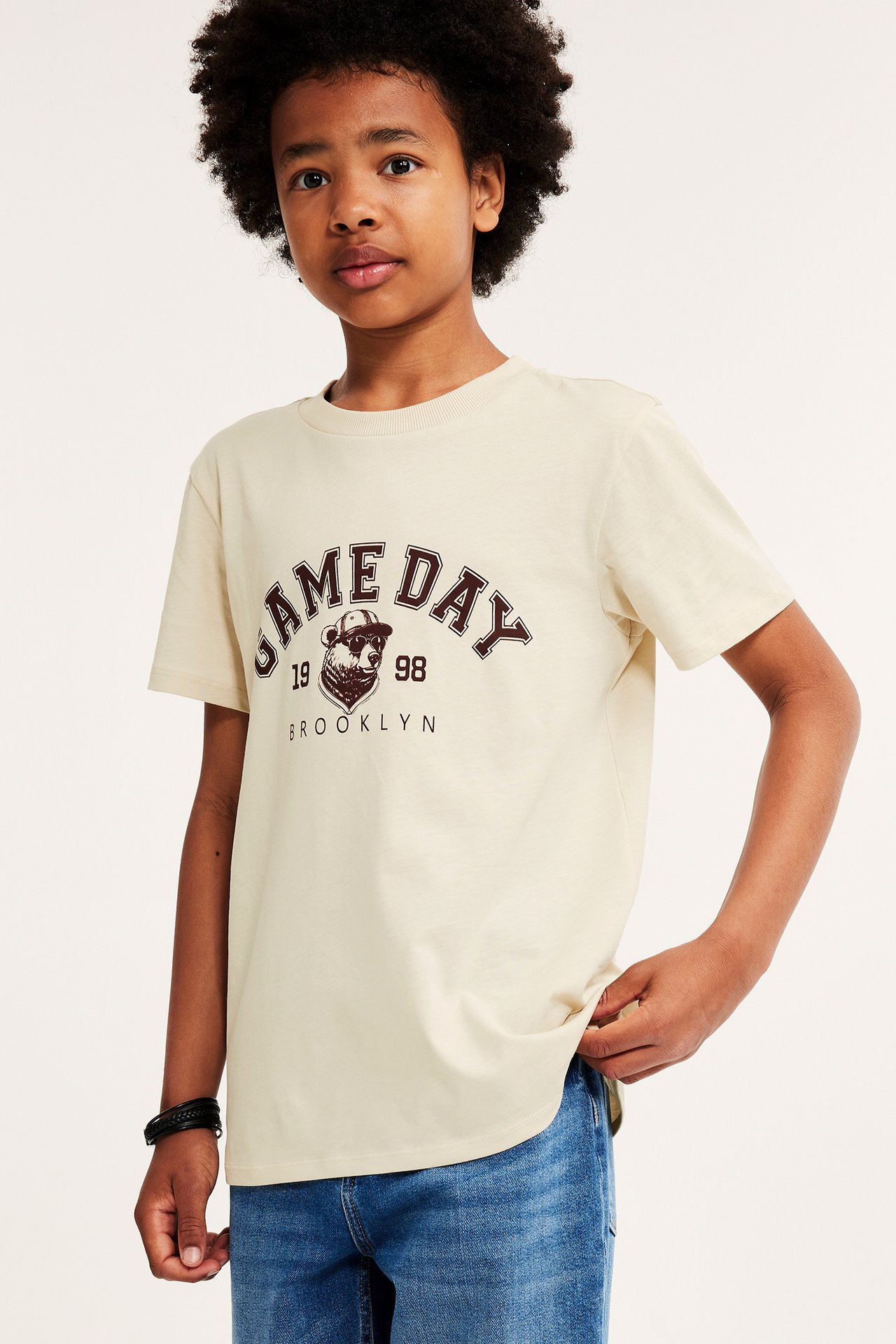 Framifrån, ett barn i beige bomulls-t-shirt med mörkbrunt tryck, blå jeans, svart armband.