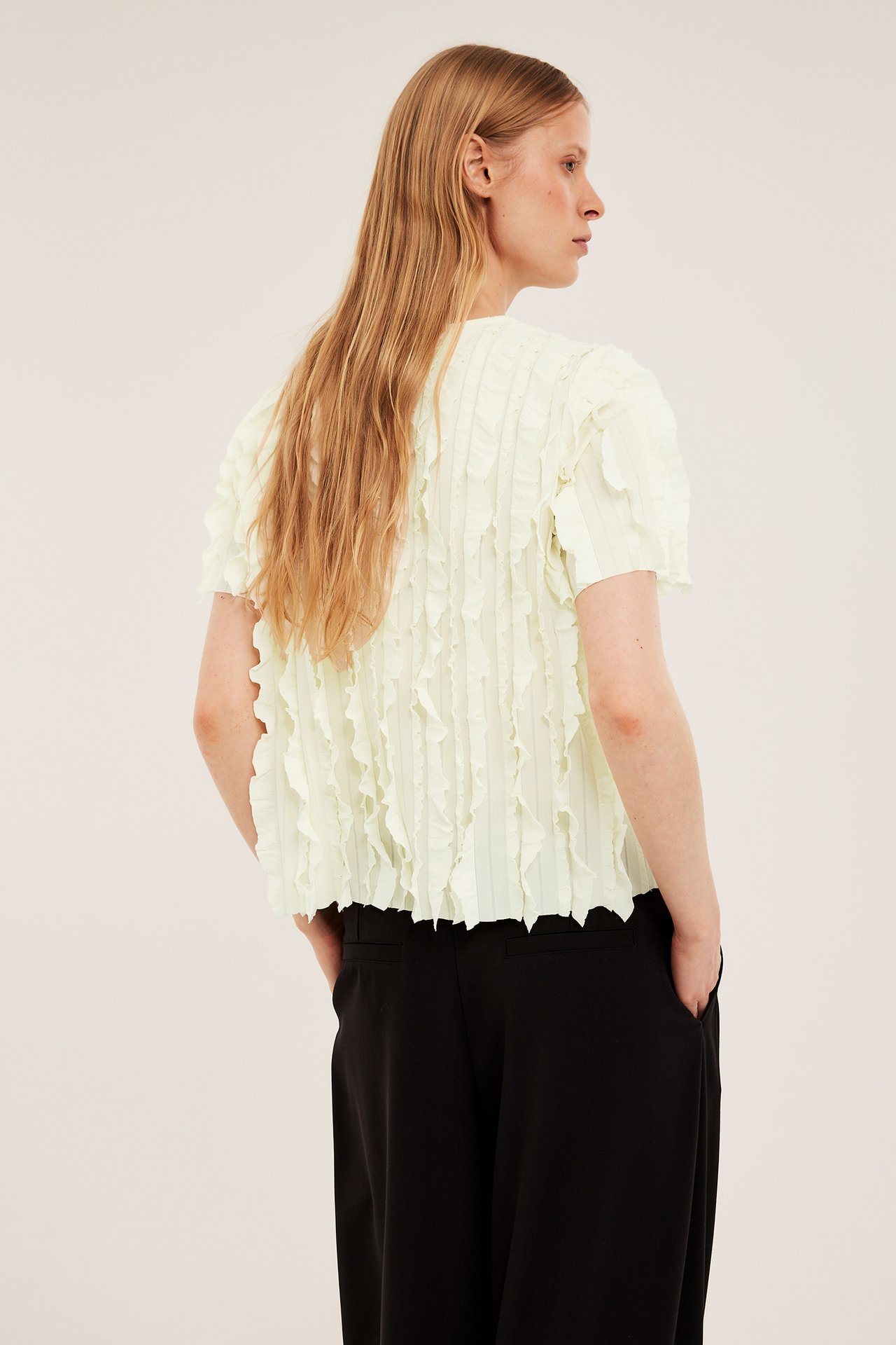 Rygg-/sidebilde av en kvinne i en offwhite strukturert rysjet topp med korte ermer og svarte bukser.