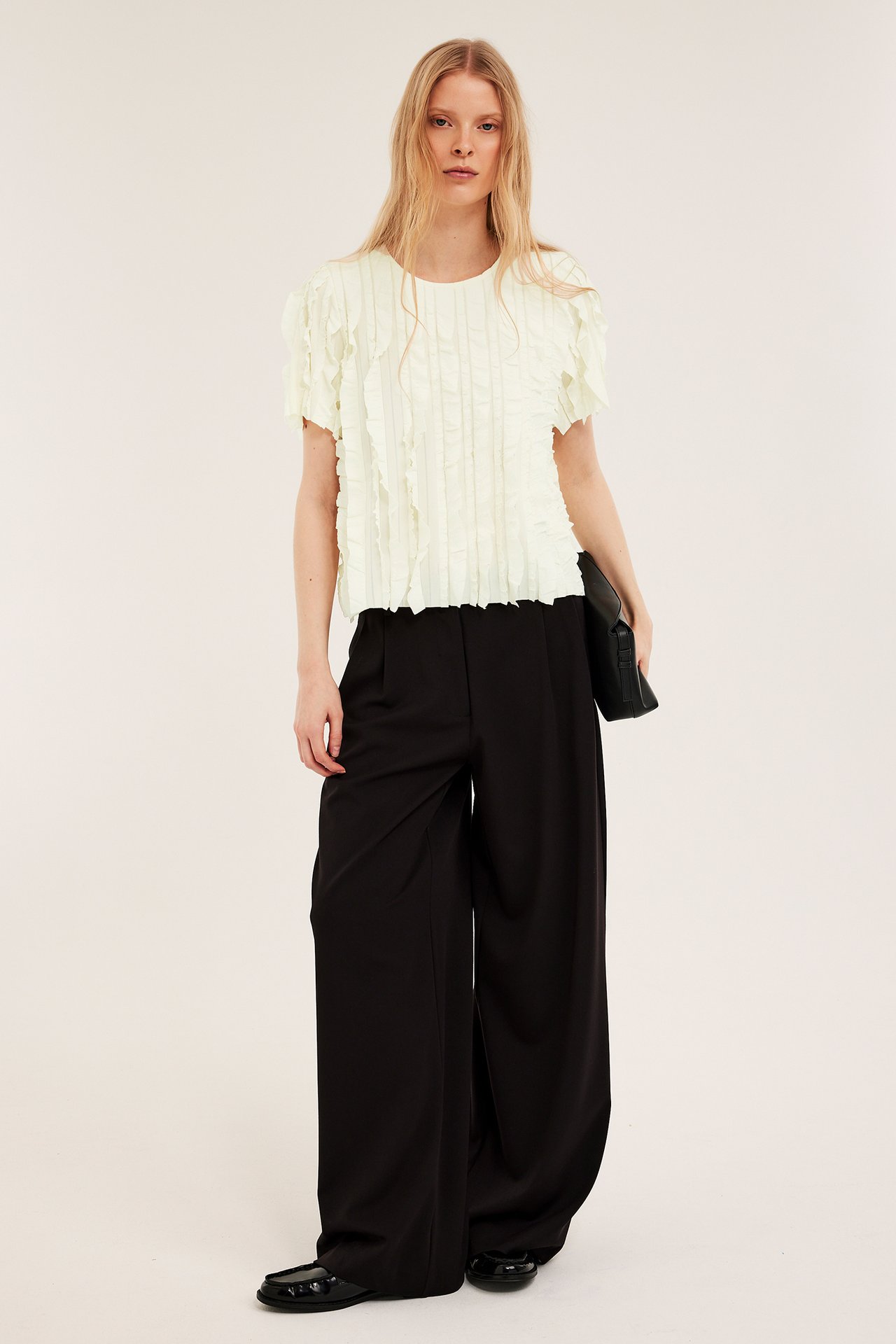 Frontutsikt: kvinne i offwhite strukturert rysjetopp, svarte vide bukser, holder svart clutch.