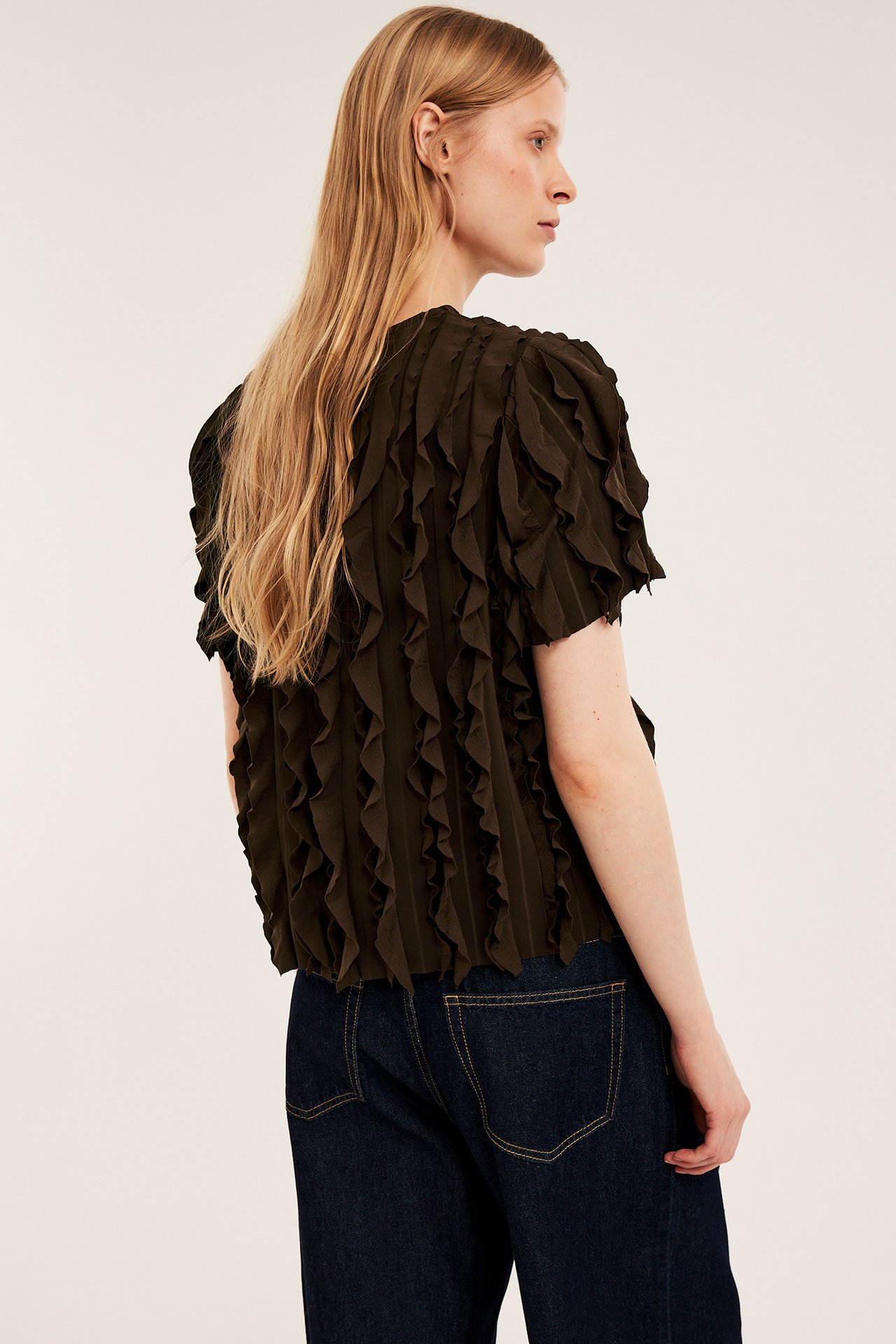 Widok z boku kobiety w ciemnobrązowym teksturowanym topie z falbanami i ciemnoniebieskich jeansach z szerokimi nogawkami.