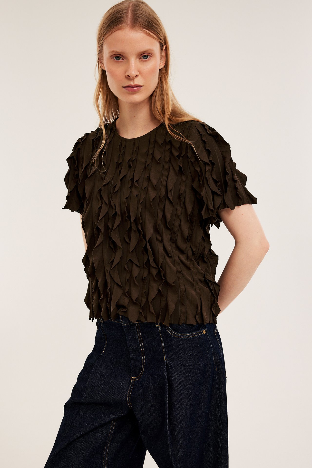 Widok z przodu: kobieta w ciemnobrązowym teksturowanym topie z falbanami i ciemnoniebieskich jeansach.