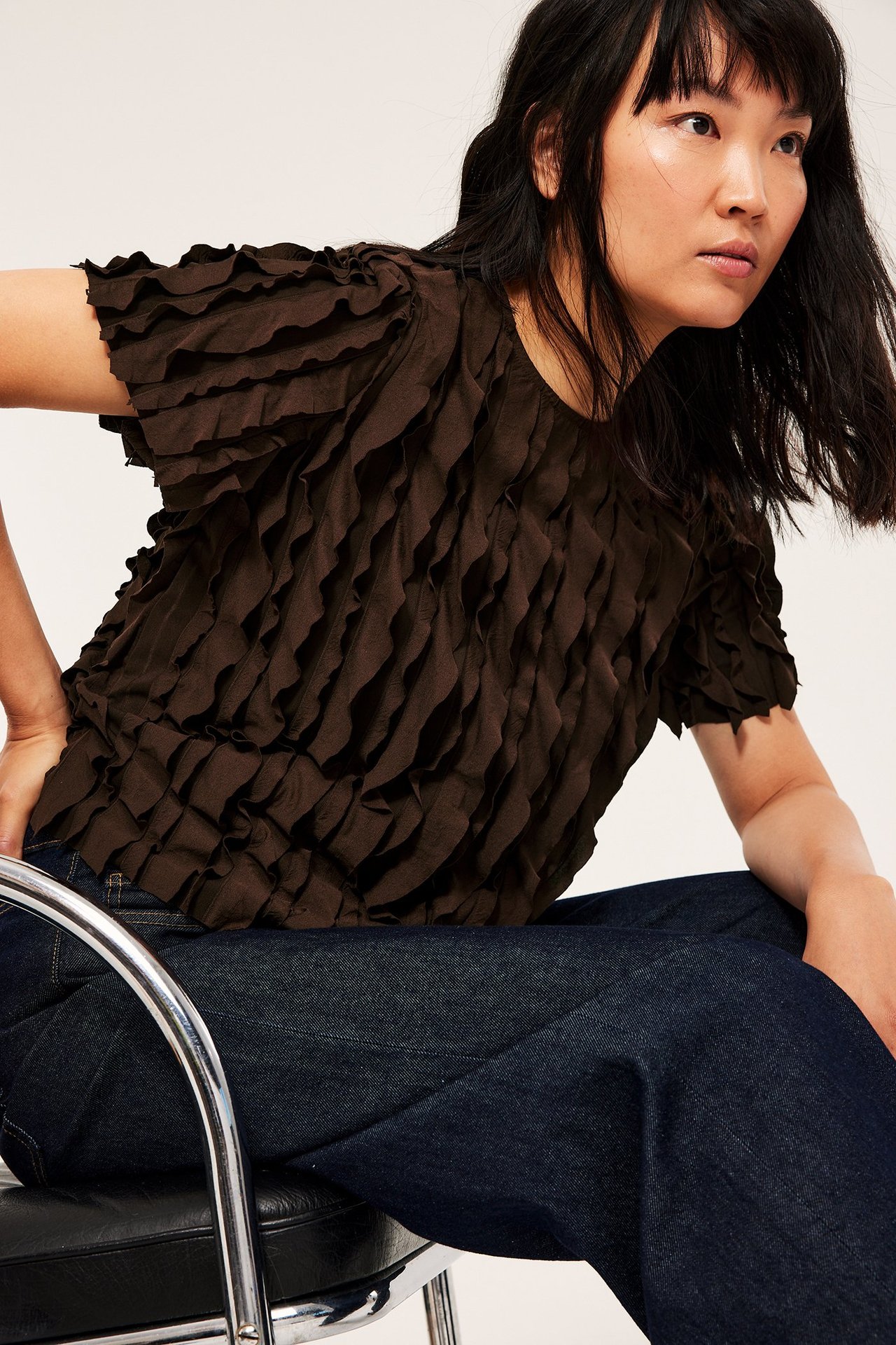 Nainen tummanruskeassa laatikkokuviossa ja volangitopissa, istuu mustalla tuolilla.