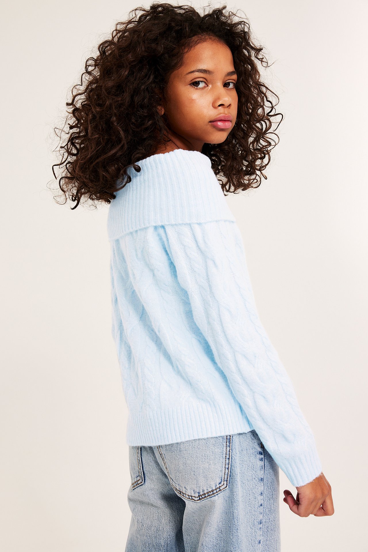 Widok z boku dziecka w jasnoniebieskim, warkoczowym swetrze off-shoulder i jasnoniebieskich jeansach.