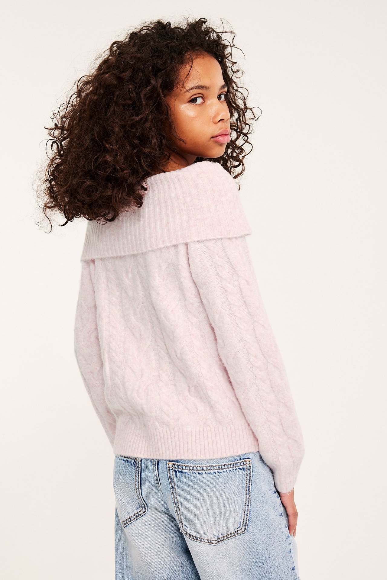 Tyttö vaaleanpunaisessa palmikkoneuleessa, olkapäät paljaana, ja vaaleansinisissä farkuissa, takaapäin olkapään yli.