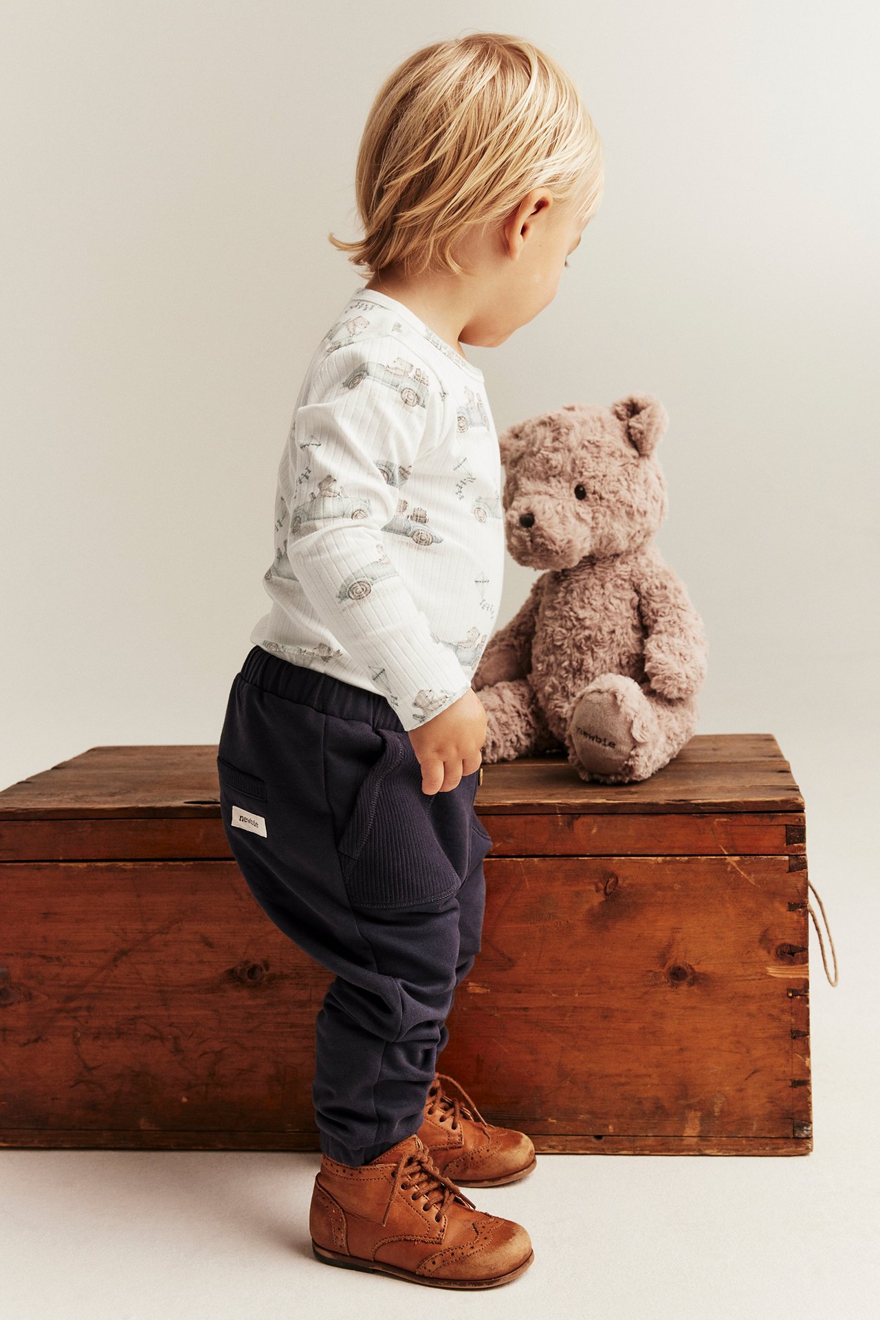 Ett barn bär en off-white ribbad body med bilmotiv, mörkblå sweatpants och bruna snörskor.