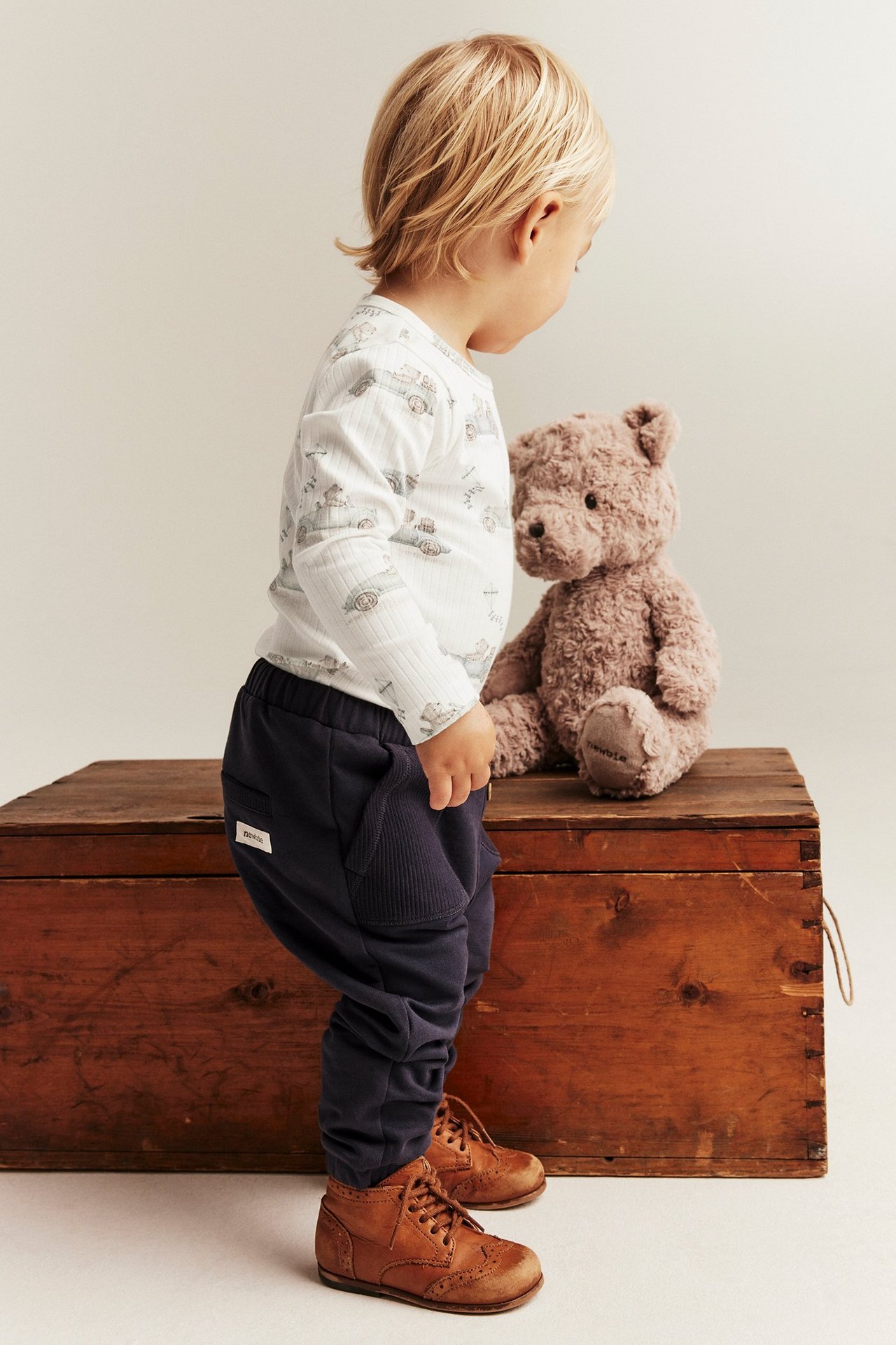 Ett barn bär en off-white ribbad body med bilmotiv, mörkblå sweatpants och bruna snörskor.