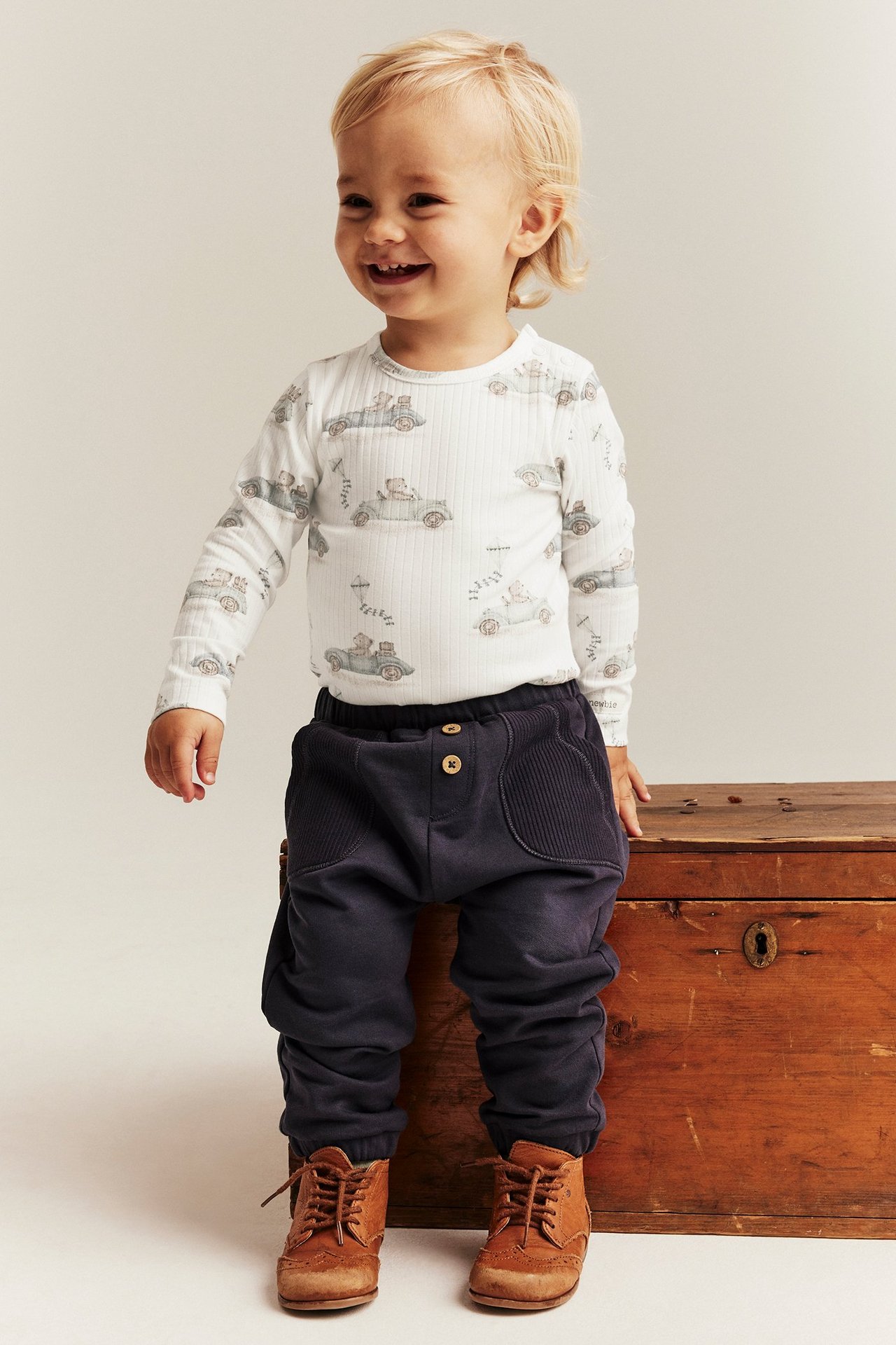 Dziecko w kremowym prążkowanym body z autem, granatowych spodniach i brązowych butach. Przód.
