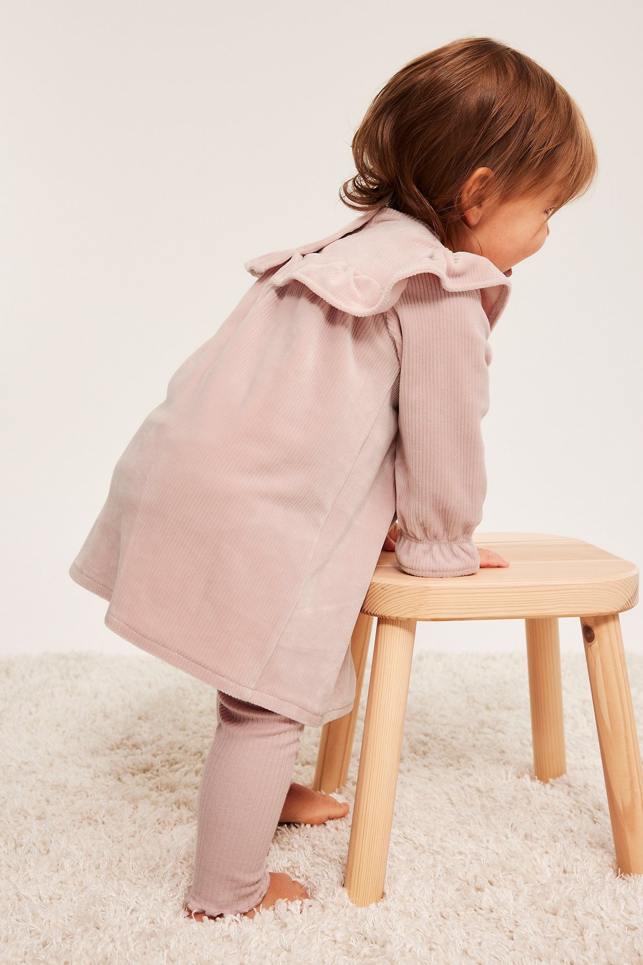 Sivukuva vauvasta vaaleanpunaisessa rypytetyssä veluurimekossa ja vaaleanpunaisessa ribbisetissä, nojaten jakkaralle.