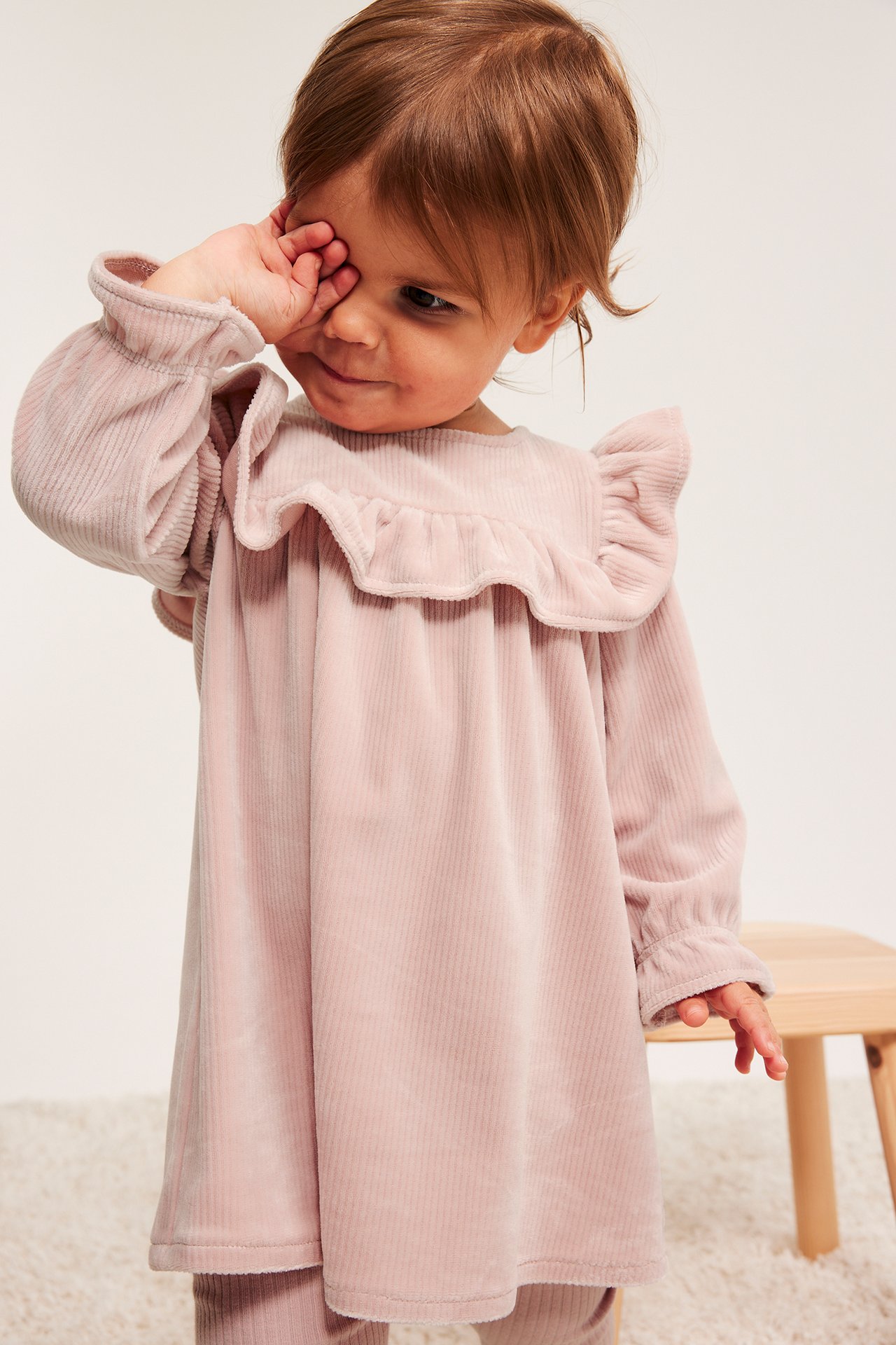 Frontvisning av en baby i en rosa ribbet velourkjole med volanger, iført matchende rosa leggings.